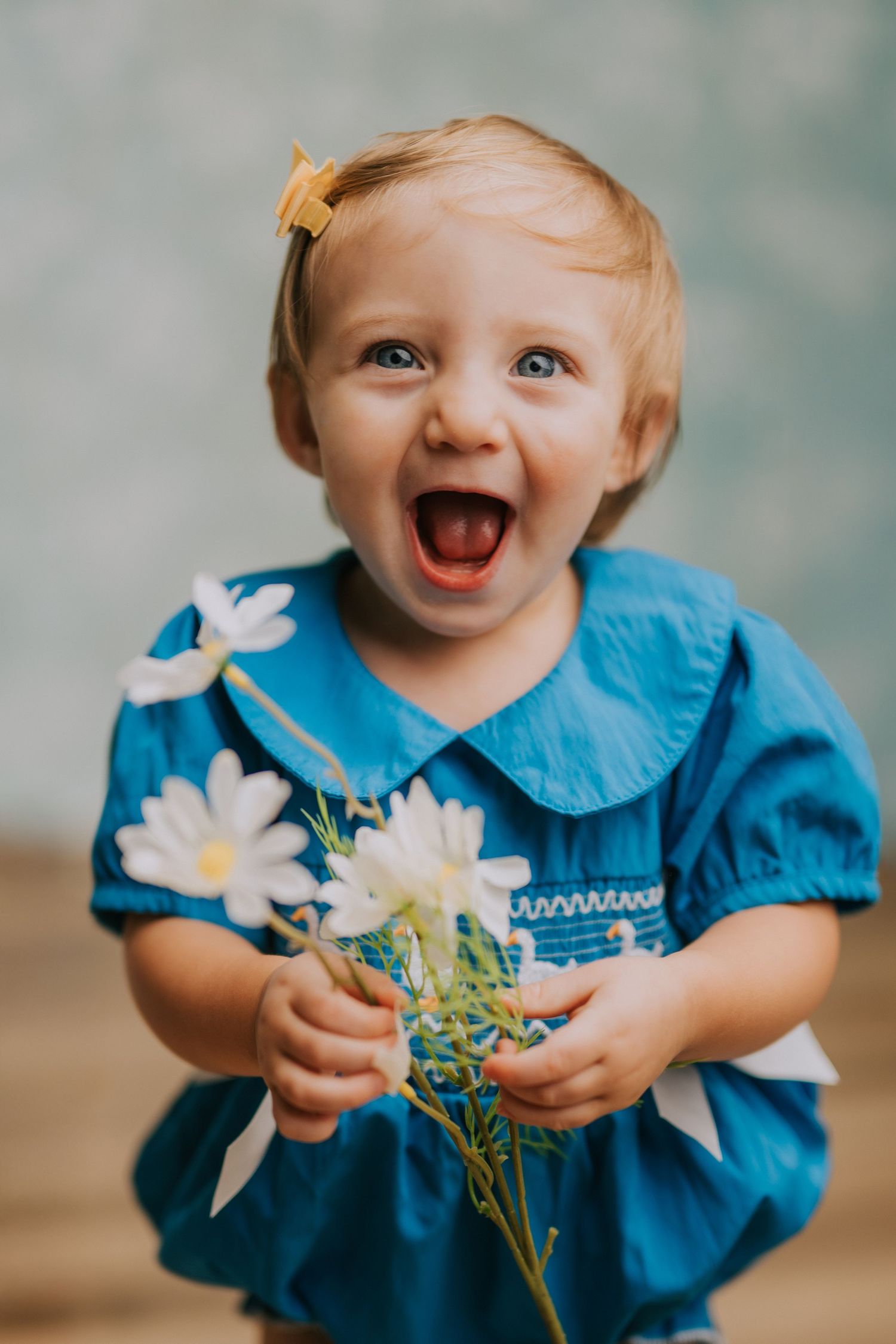 Baby in blue dress holding white flowers and showing joyful expressions.