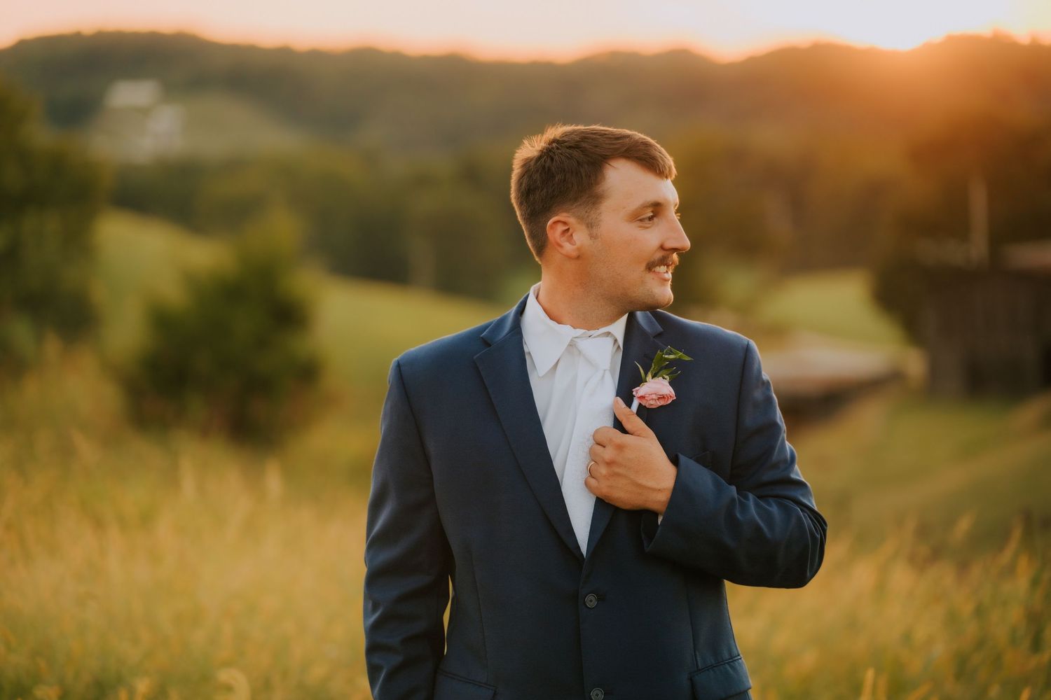 Groom in navy suit adjusting tie during golden hour outdoor photoshoot.