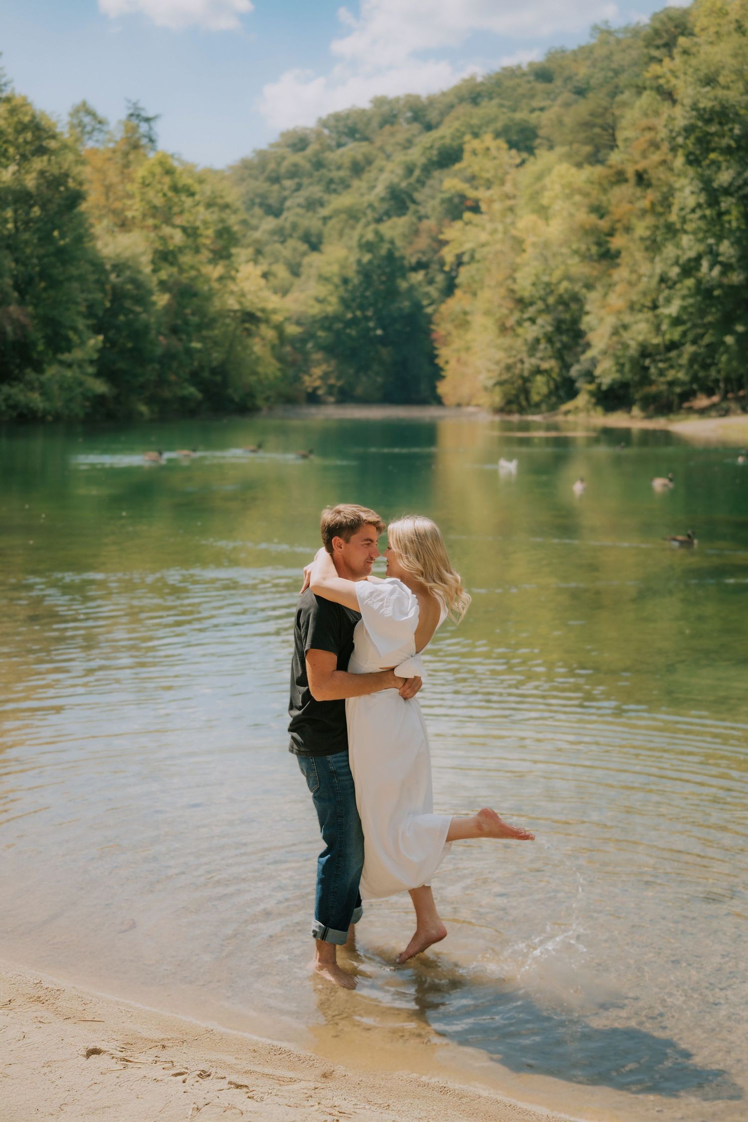 Couple embracing in shallow lake water surrounded by lush summer forest landscape.