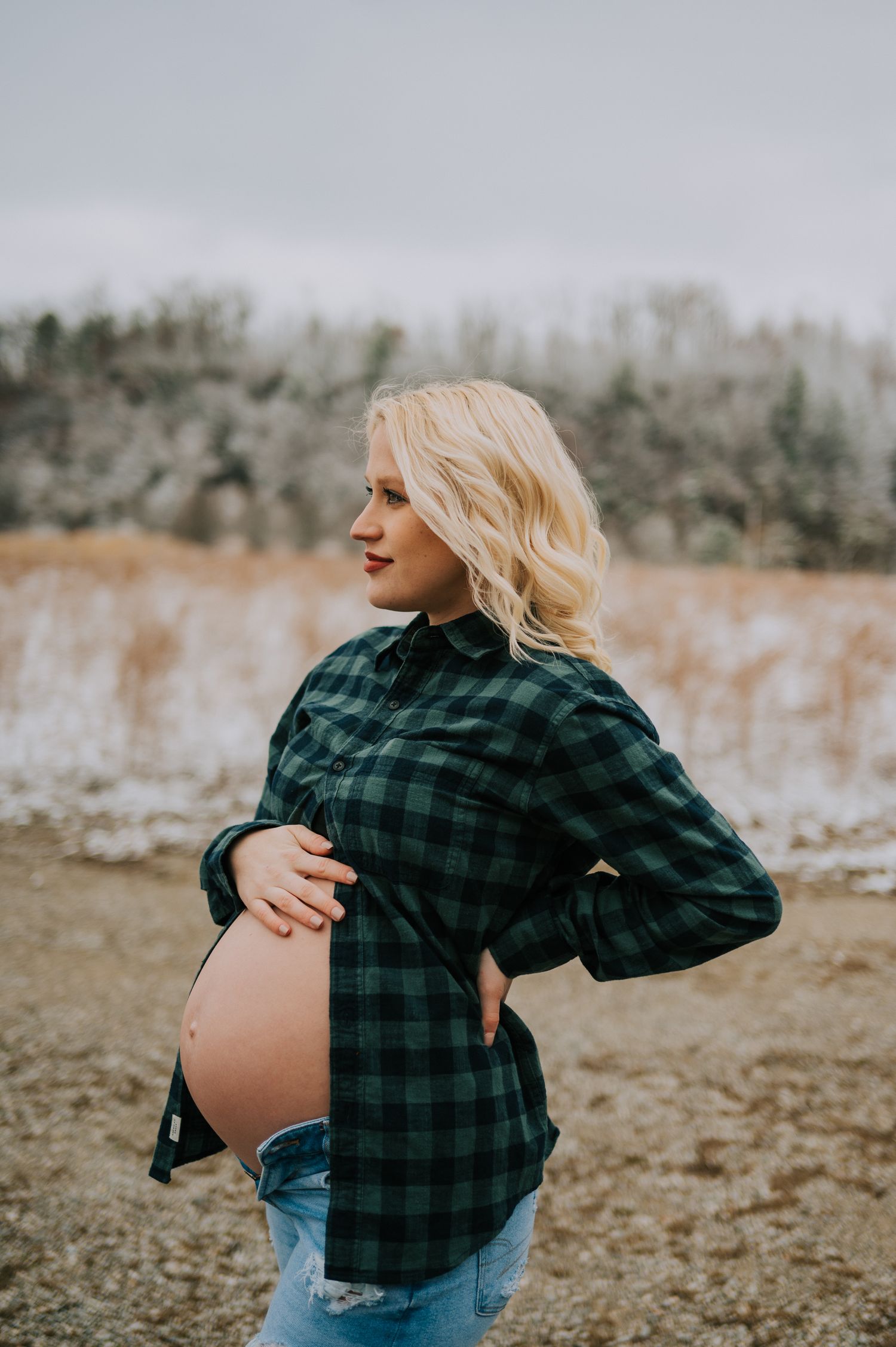 Pregnant woman in plaid shirt showing baby bump against winter landscape.