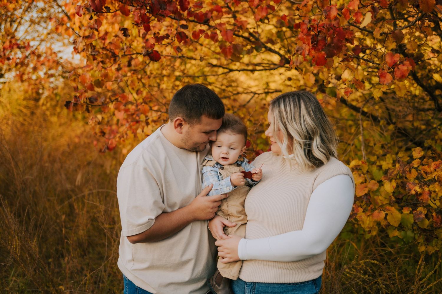 Couple holding small child against backdrop of golden fall foliage at sunset.