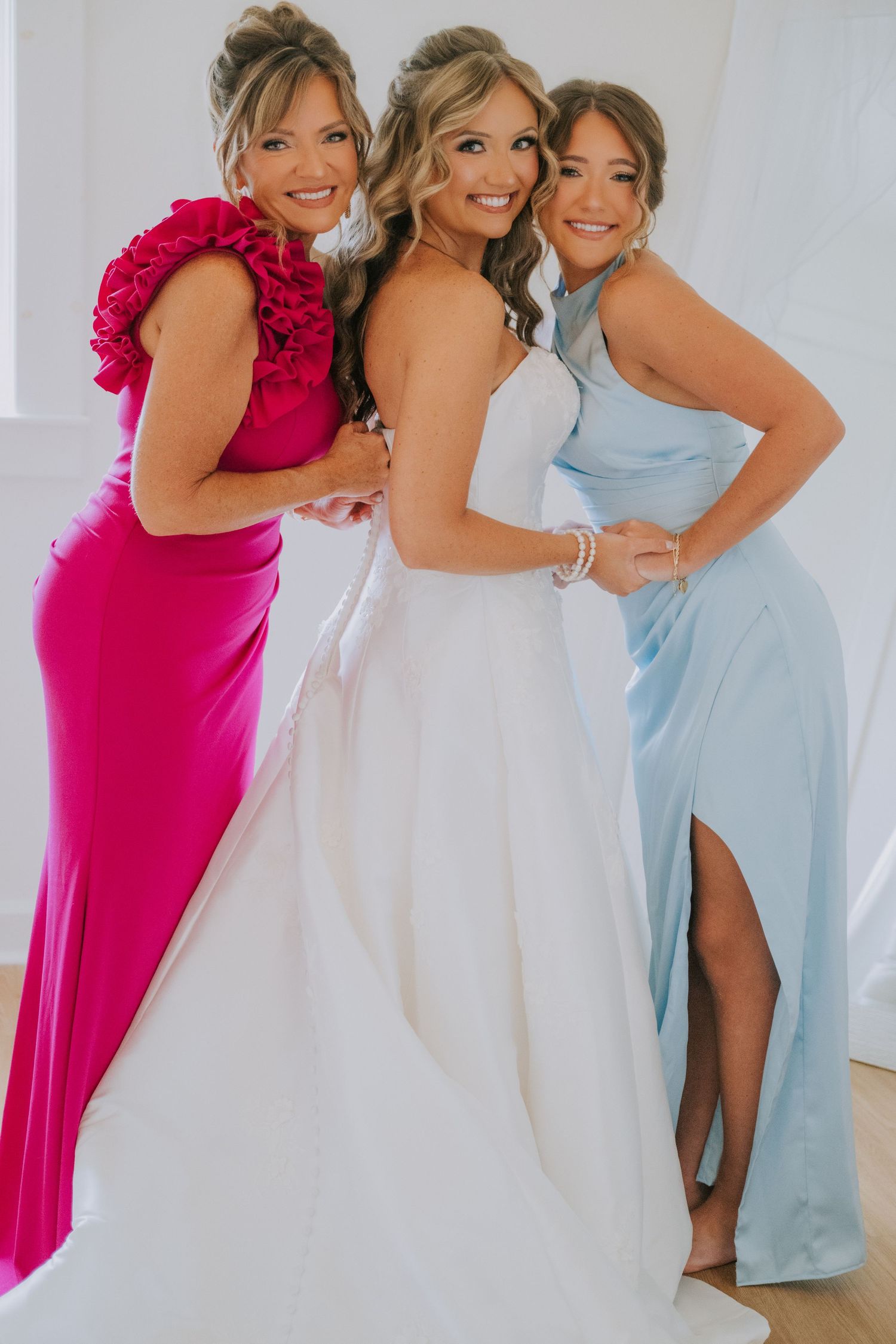 Bride in white gown with two bridesmaids in pink and blue dresses posing together at a wedding.