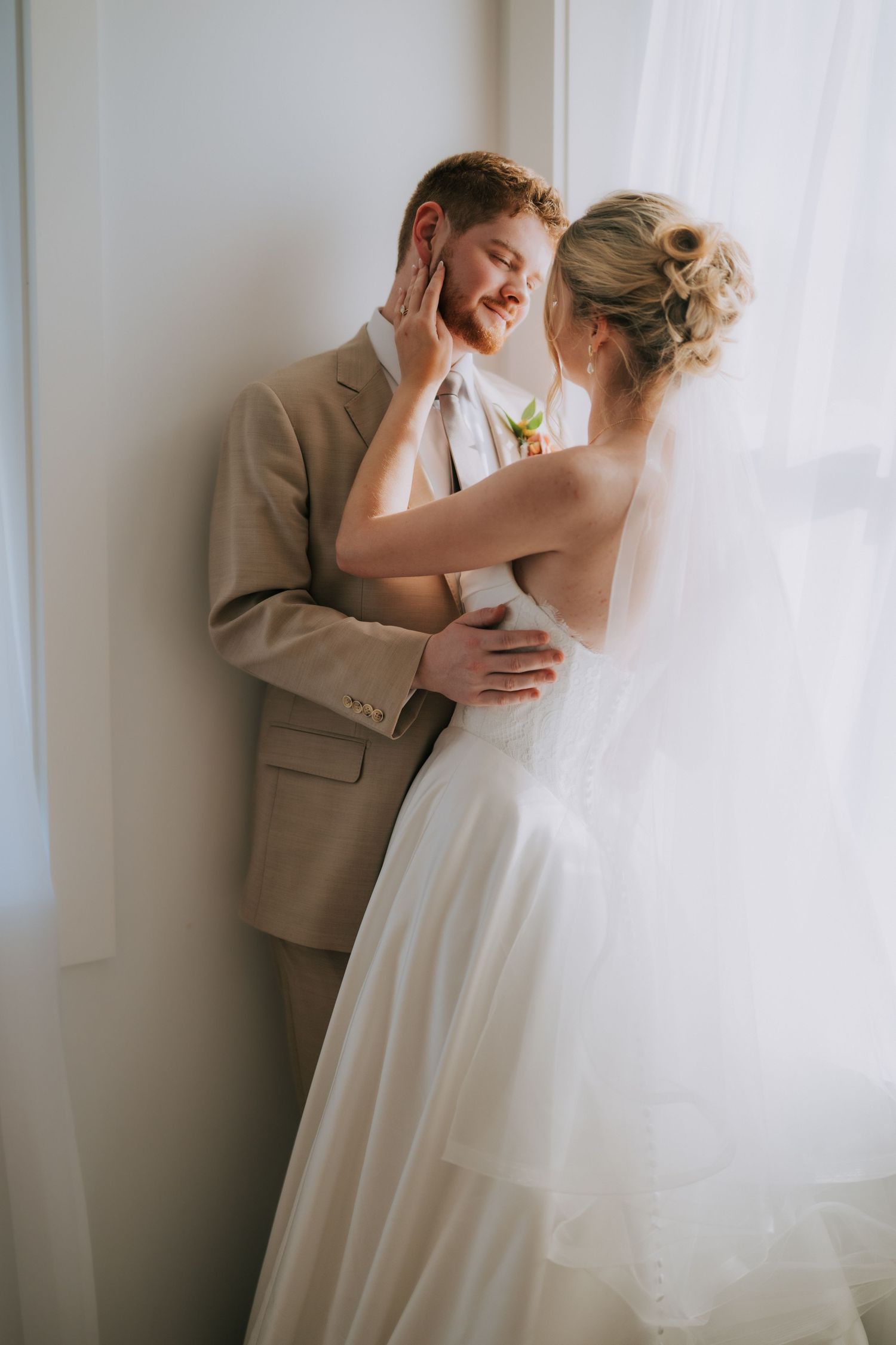 Bride and groom share a romantic moment by a window, with bride in white gown touching grooms face in a tan suit.