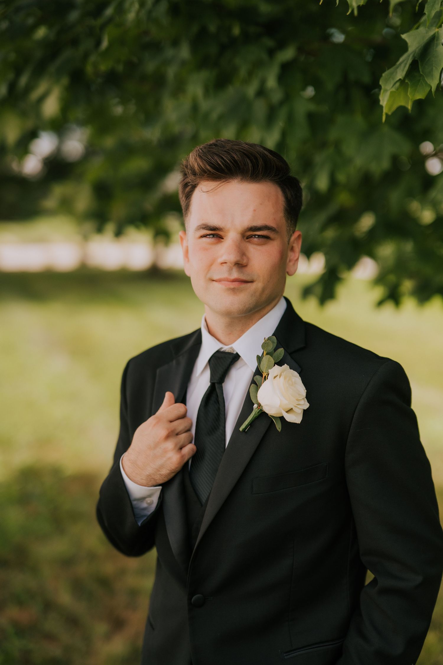 A groom adjusts his tie while wearing a classic black tuxedo and white boutonniere.