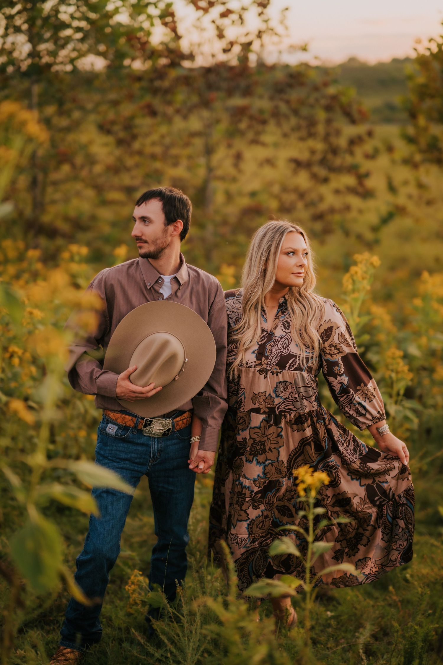 Couple walks through wildflower field at sunset in western attire.