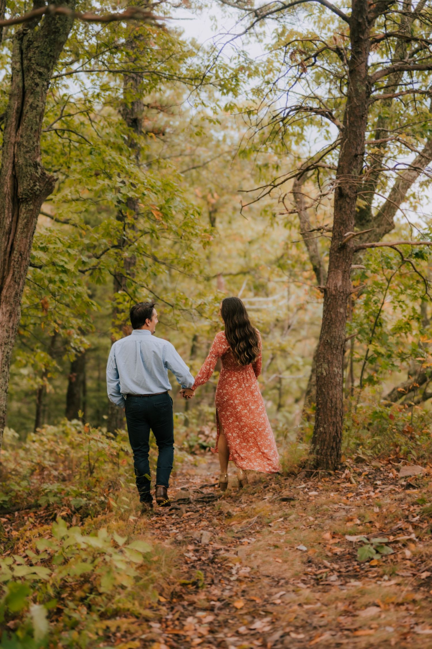 A couple walks hand in hand down a leaf-covered forest trail in autumn.