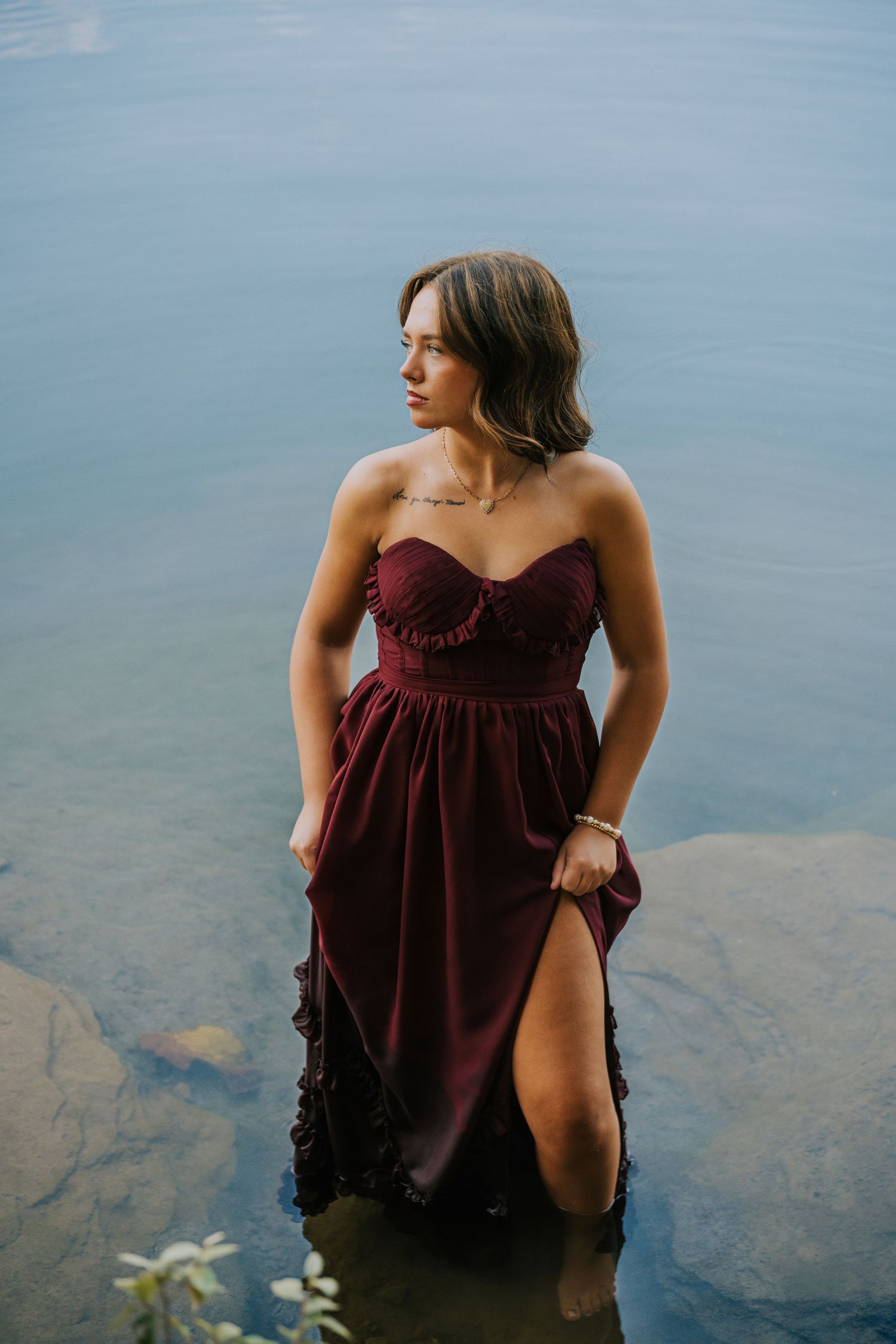 Person in burgundy dress sitting by calm water's edge during a photo session.