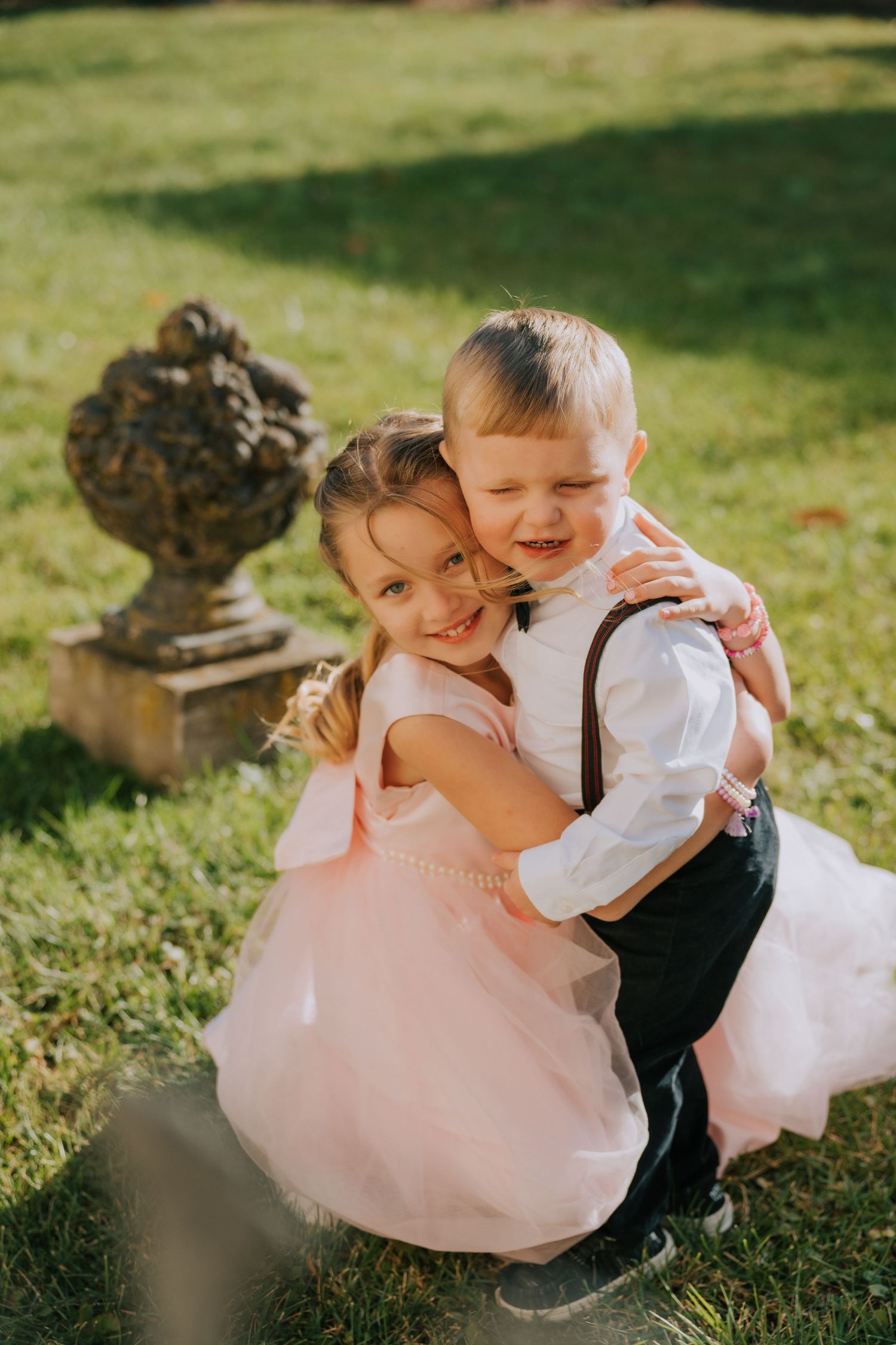 A flower girl in pink and ring bearer in suspenders share a sweet moment on the lawn by a garden ornament.