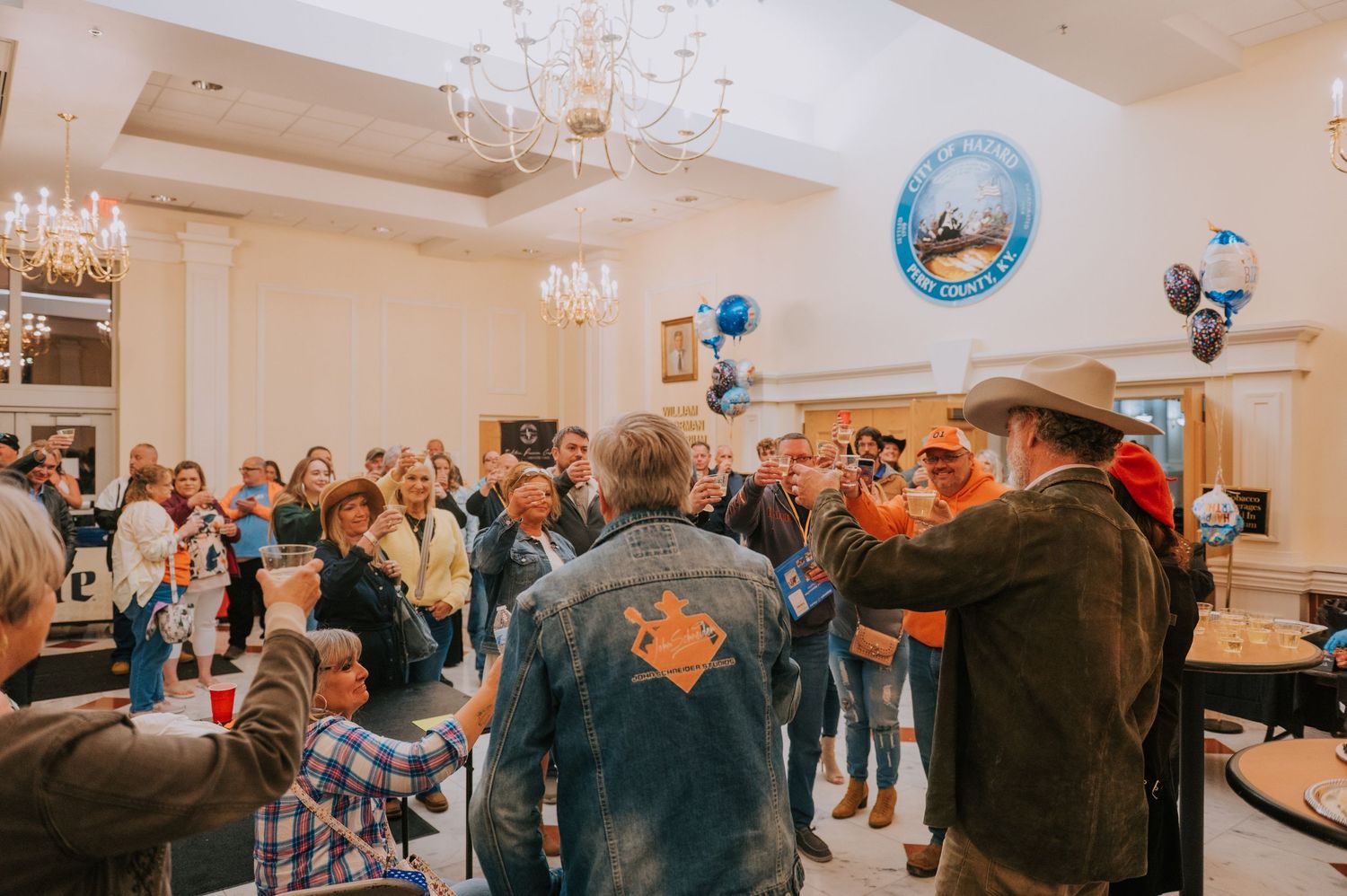A crowd of people gathering in an elegant ballroom with chandeliers and festive blue balloons for a celebration event.
