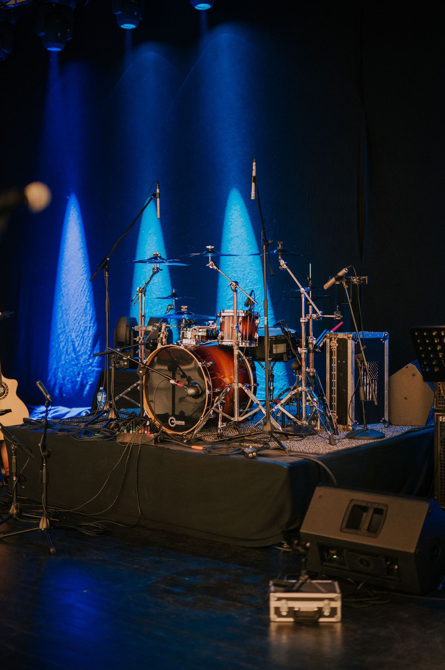 Drum kit and musical equipment set up on dark stage with dramatic blue spotlights illuminating the performance space.