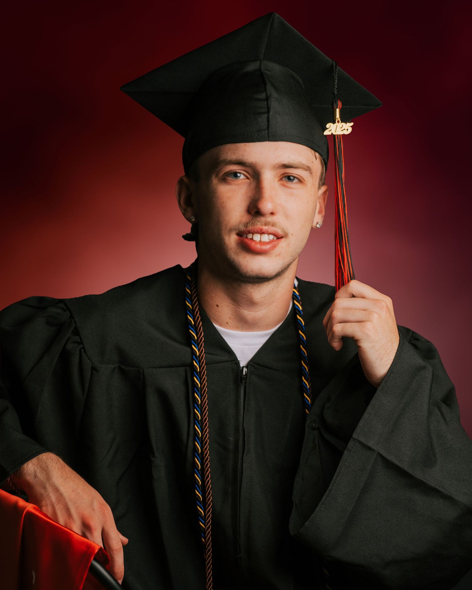 Young man in black graduation cap and gown holding tassel, posing for senior portrait against red background.