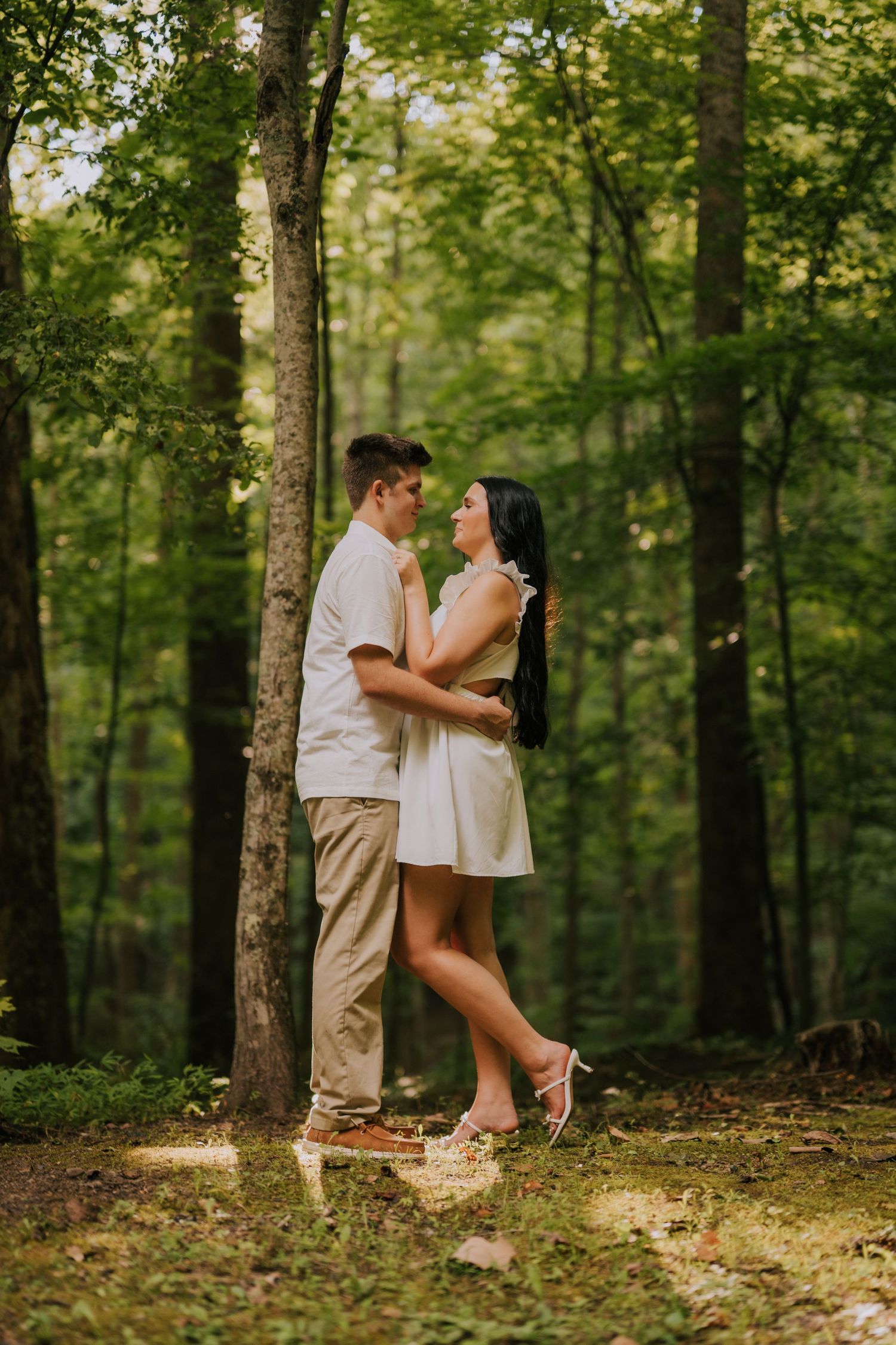 A couple in white outfits shares a romantic moment among tall trees in a forest setting.