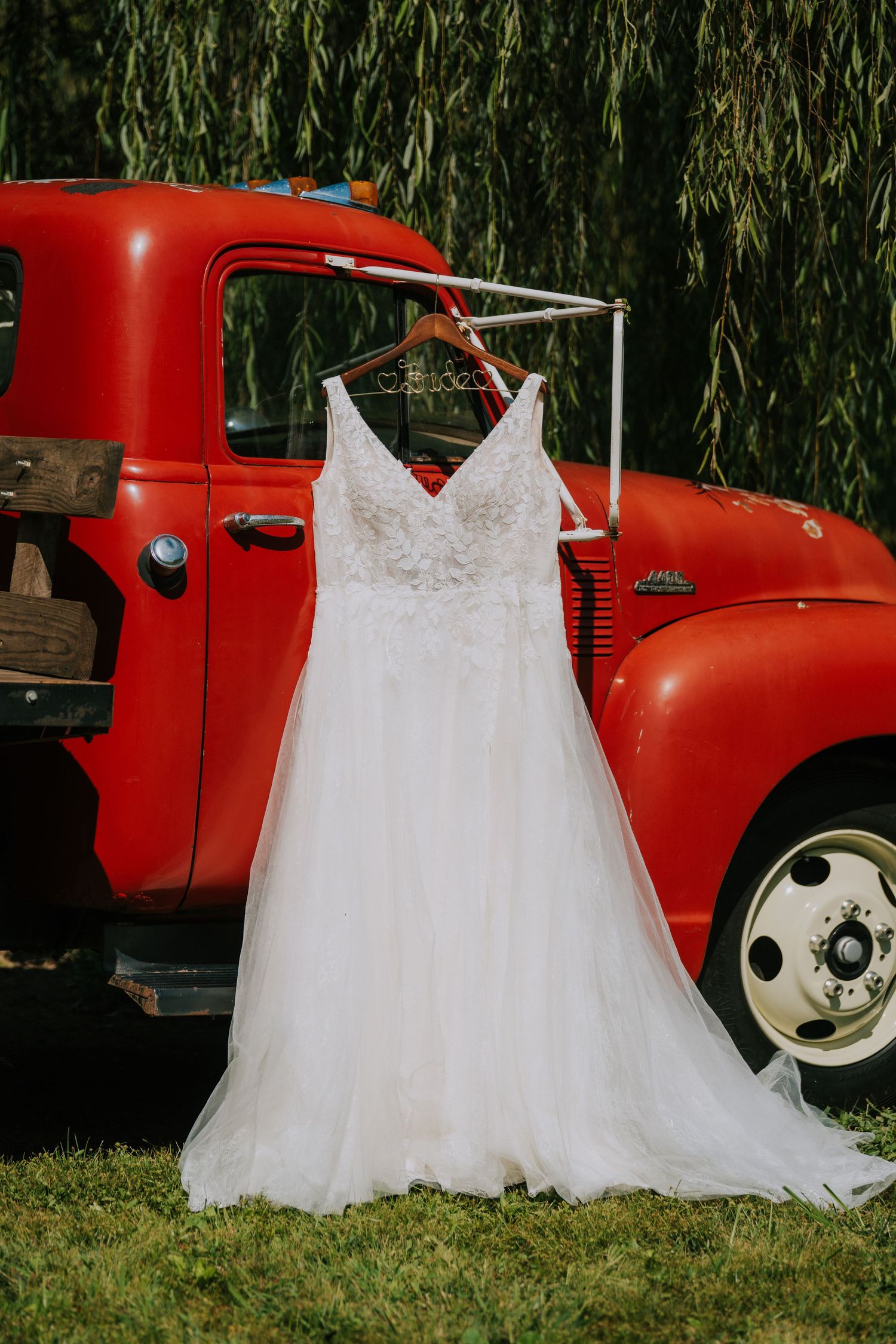A white lace wedding dress hangs displayed against a vintage red pickup truck under willow trees.