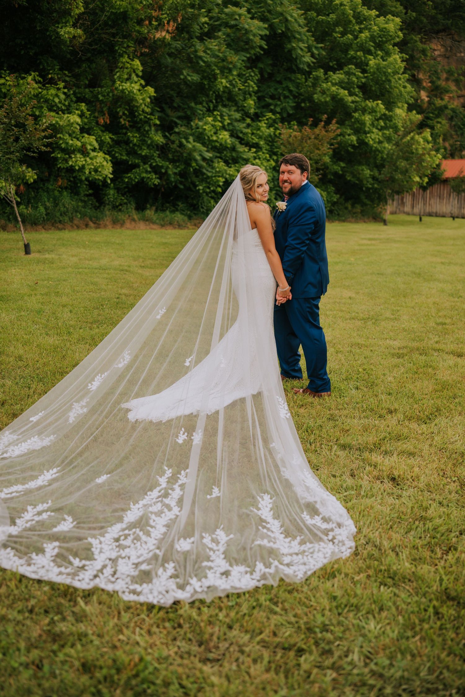 Intricate lace wedding veil trails behind on green grass during outdoor photo session.