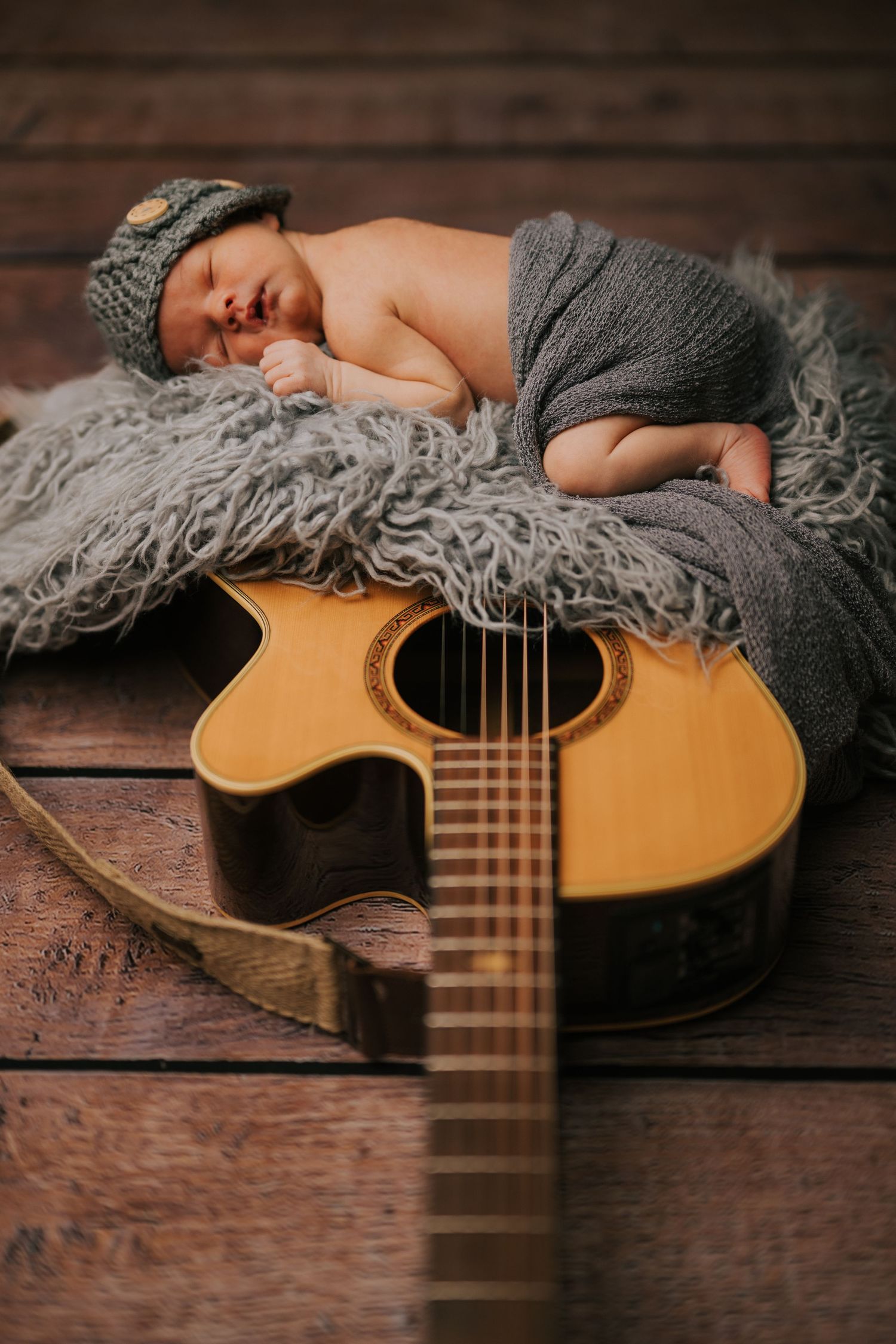Newborn photography setup with acoustic guitar and gray blanket on wooden floor.