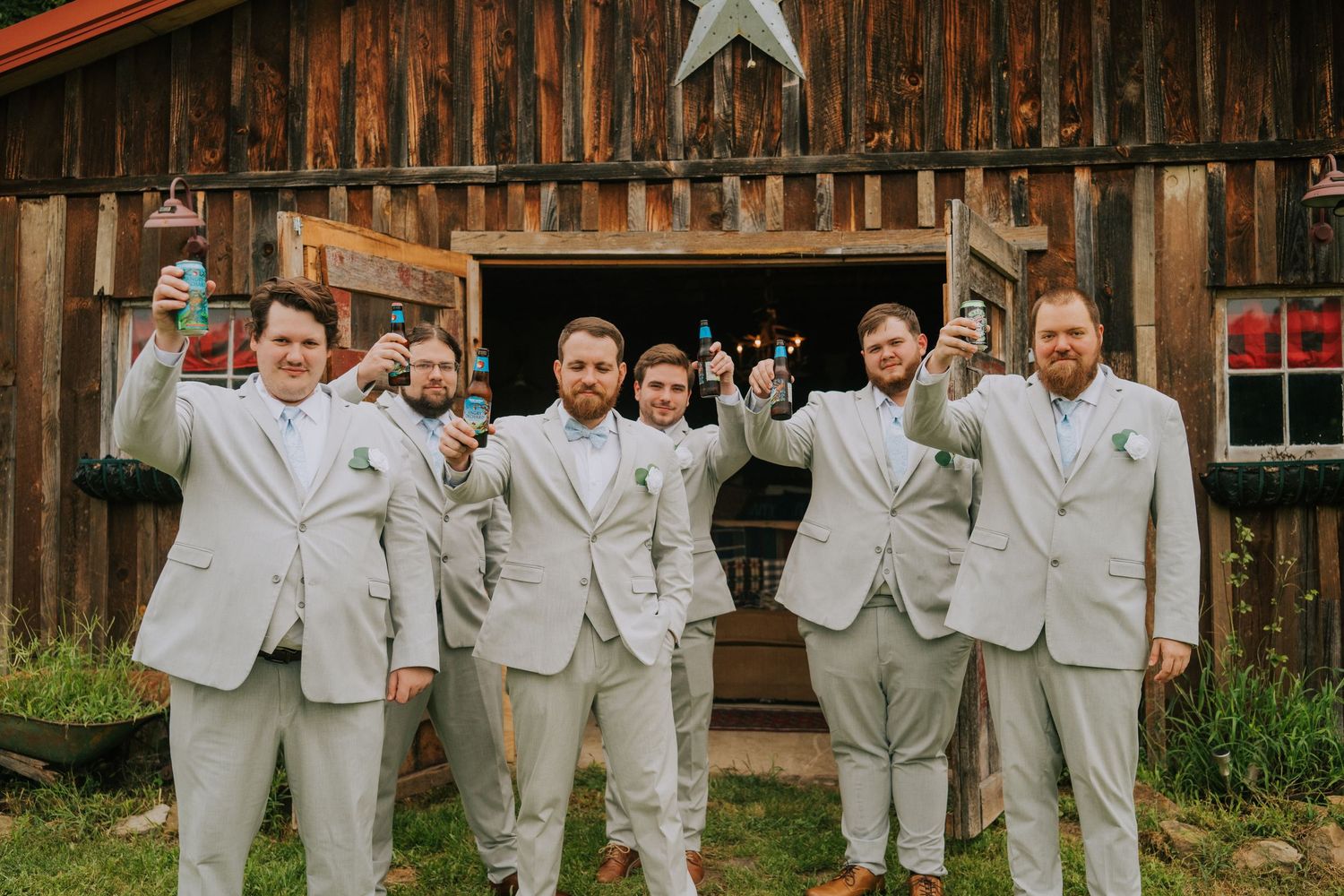 Groomsmen in light gray suits pose playfully in front of a rustic barn backdrop.
