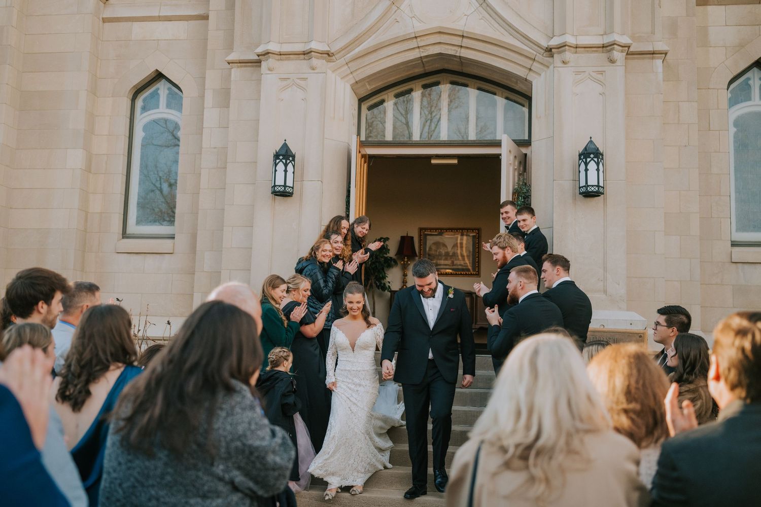 Wedding party exits church as guests celebrate on the stone steps outside.