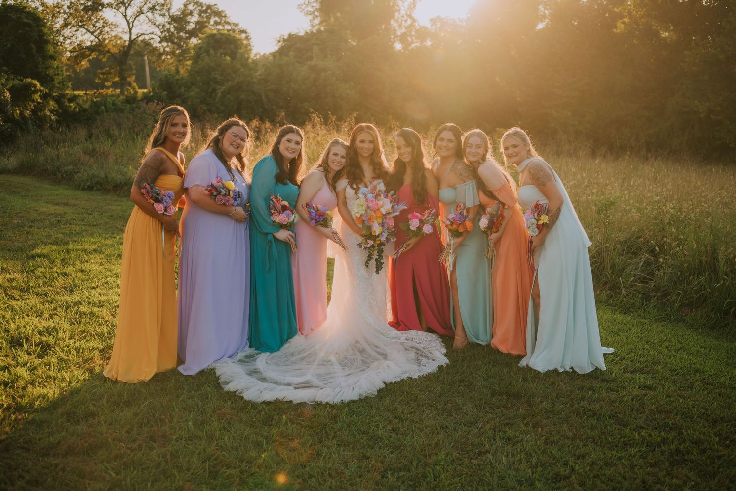 Wedding party in colorful dresses stands in a grassy field at sunset.