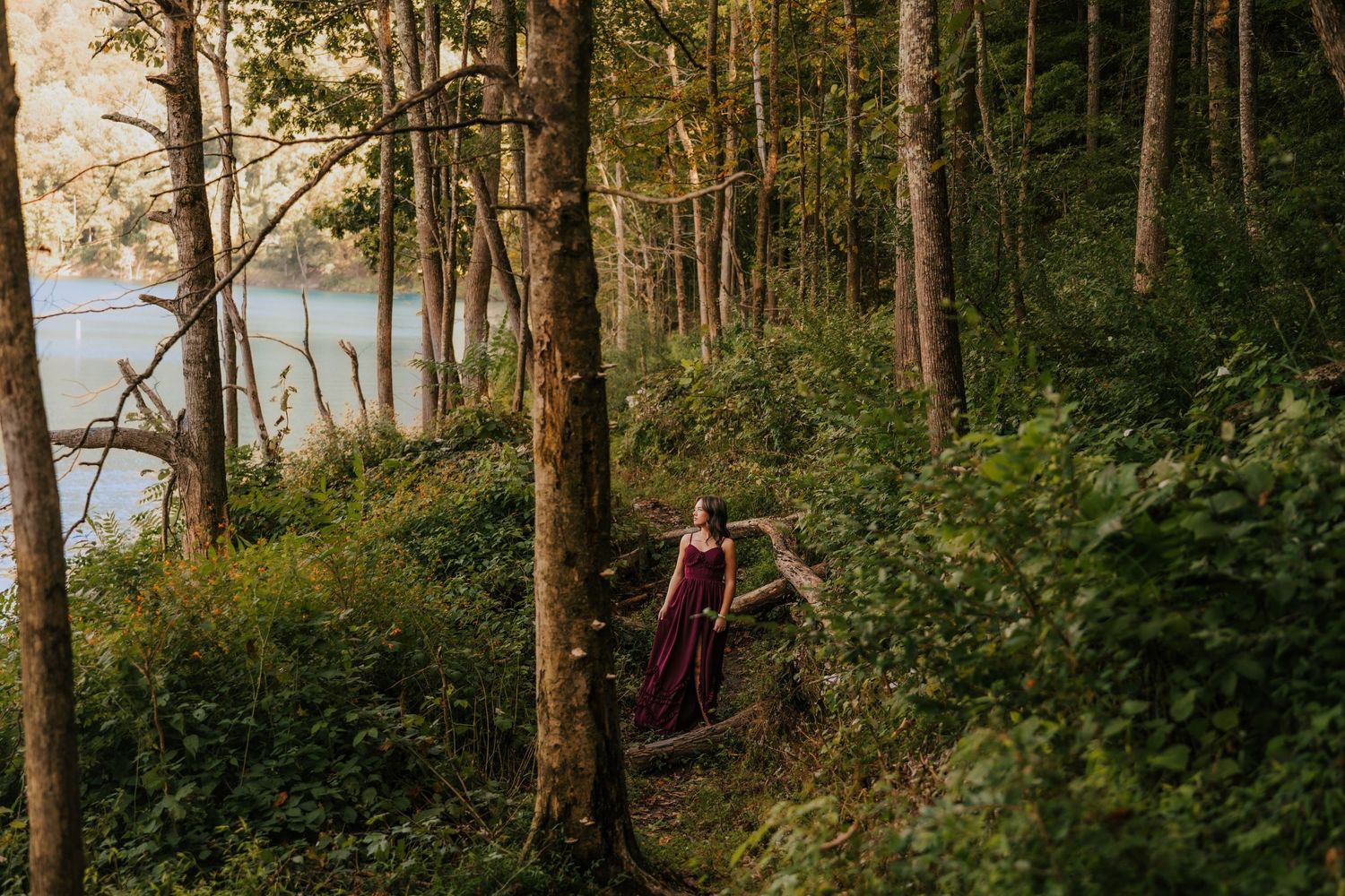 A figure in a flowing dress walks through a wooded area near a serene lake.