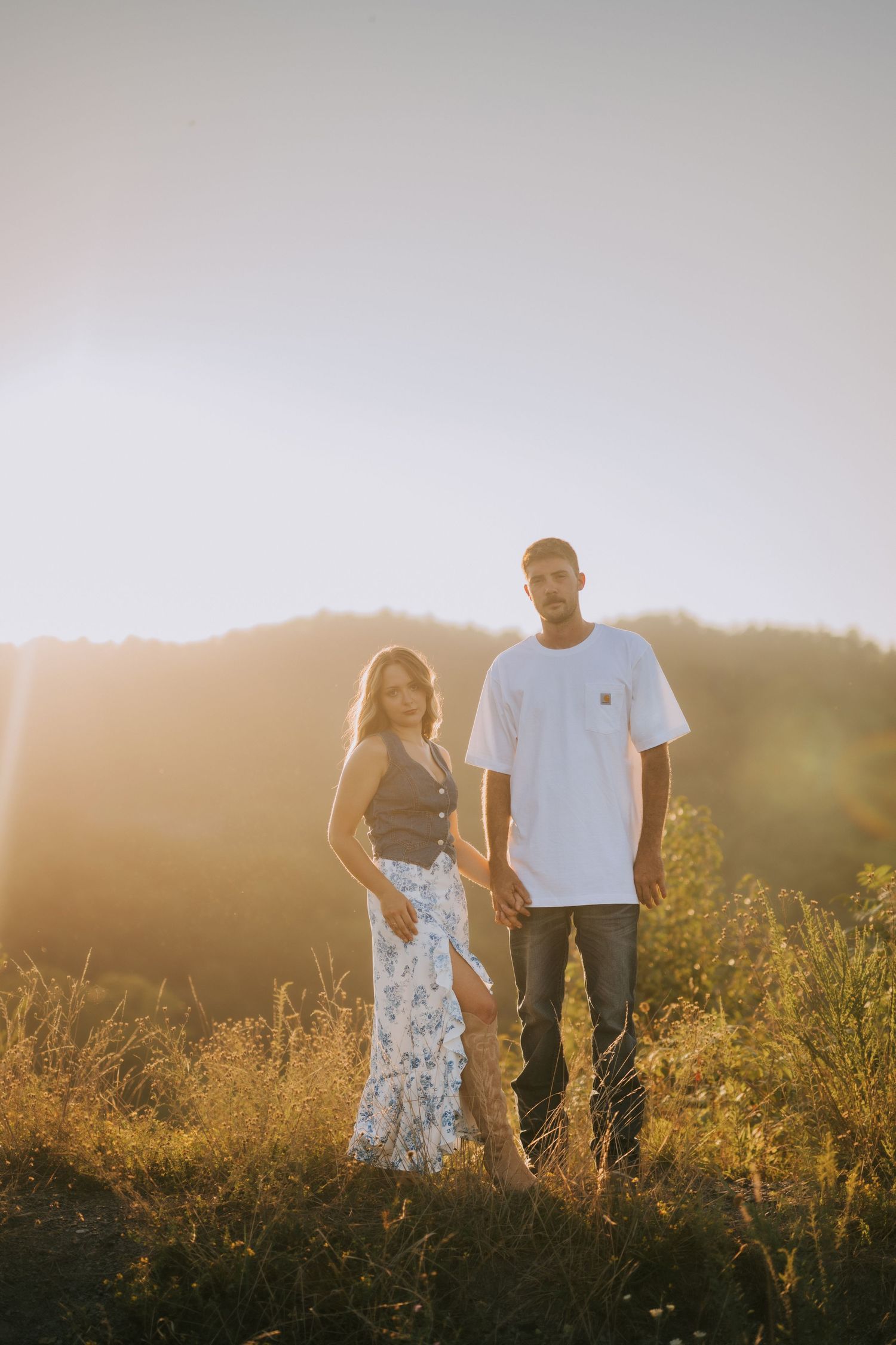 A couple stands together in a sunlit meadow during golden hour surrounded by tall grass.