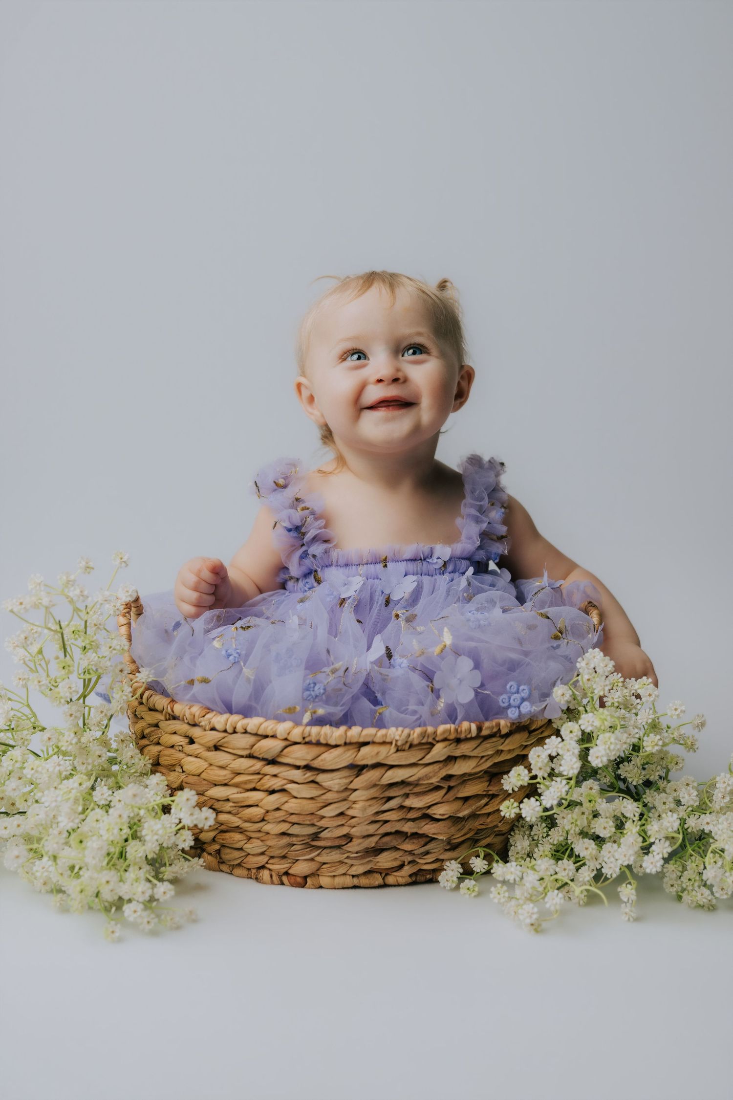 Smiling baby girl in purple tulle dress sitting in wicker basket surrounded by white flowers on gray background.