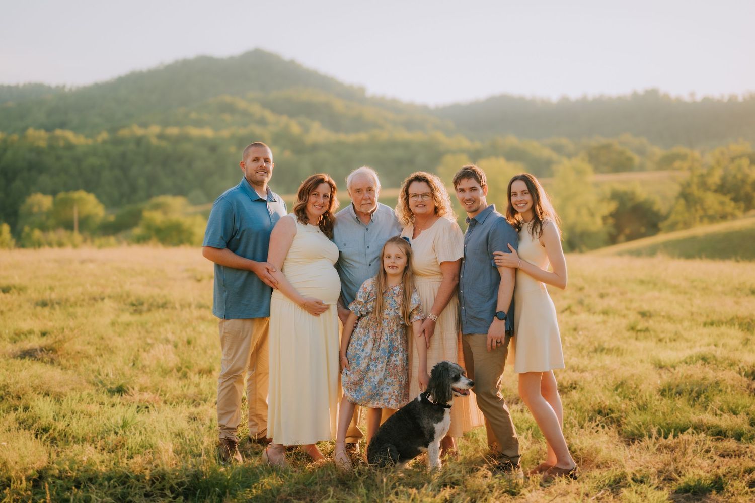 Family of seven poses with a black dog in a sunny green field with rolling hills in the background.