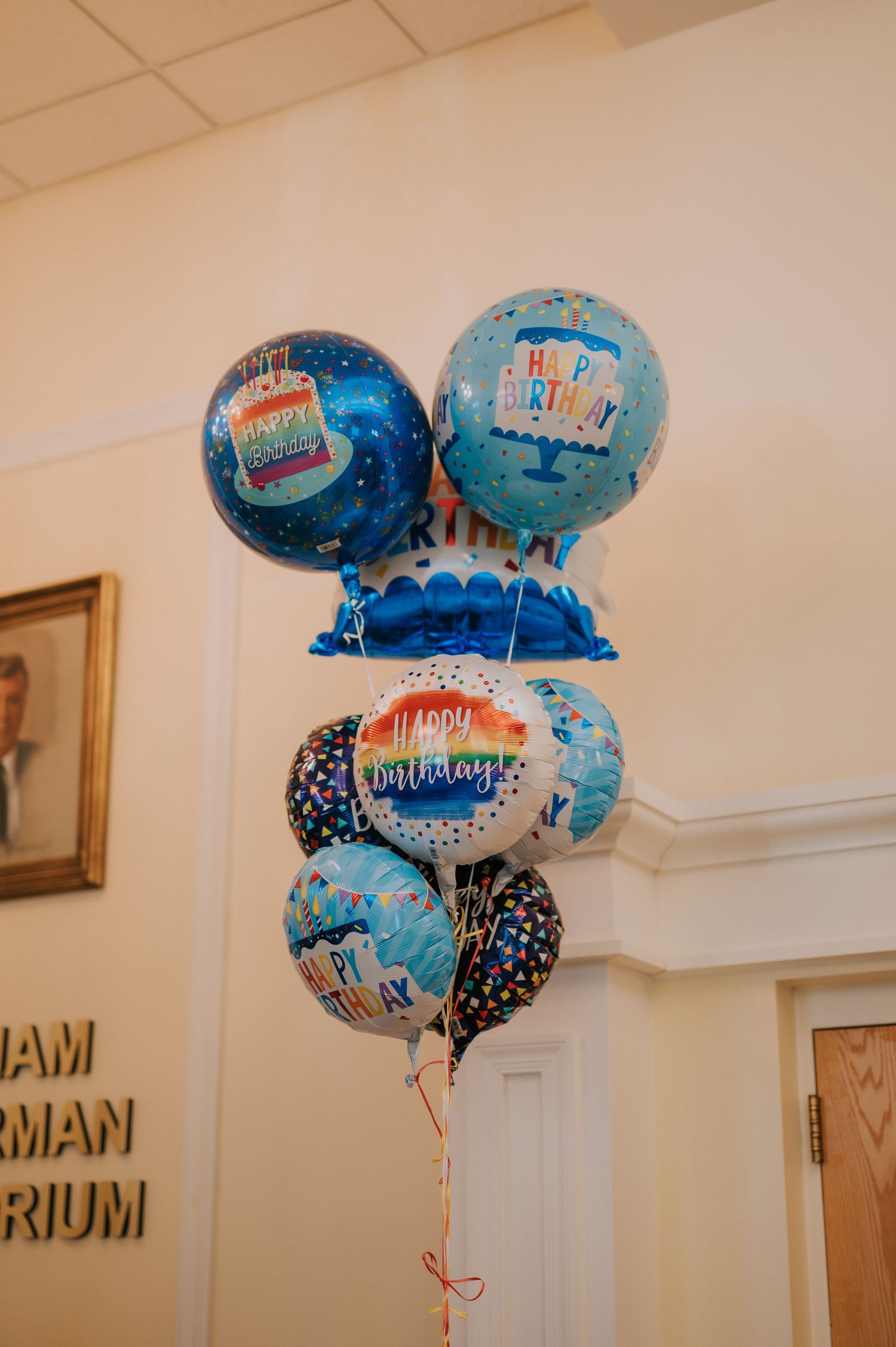 A cluster of blue and white birthday balloons tied together floating in an indoor hallway near a door.