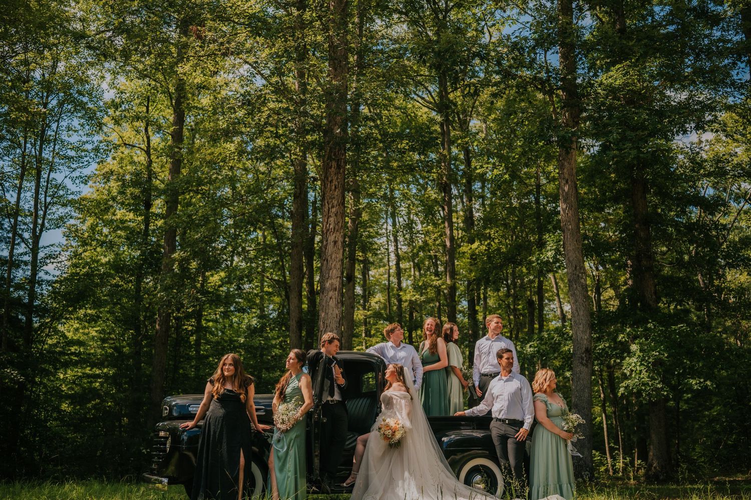 Wedding party poses together in front of vintage car in sunlit forest setting.