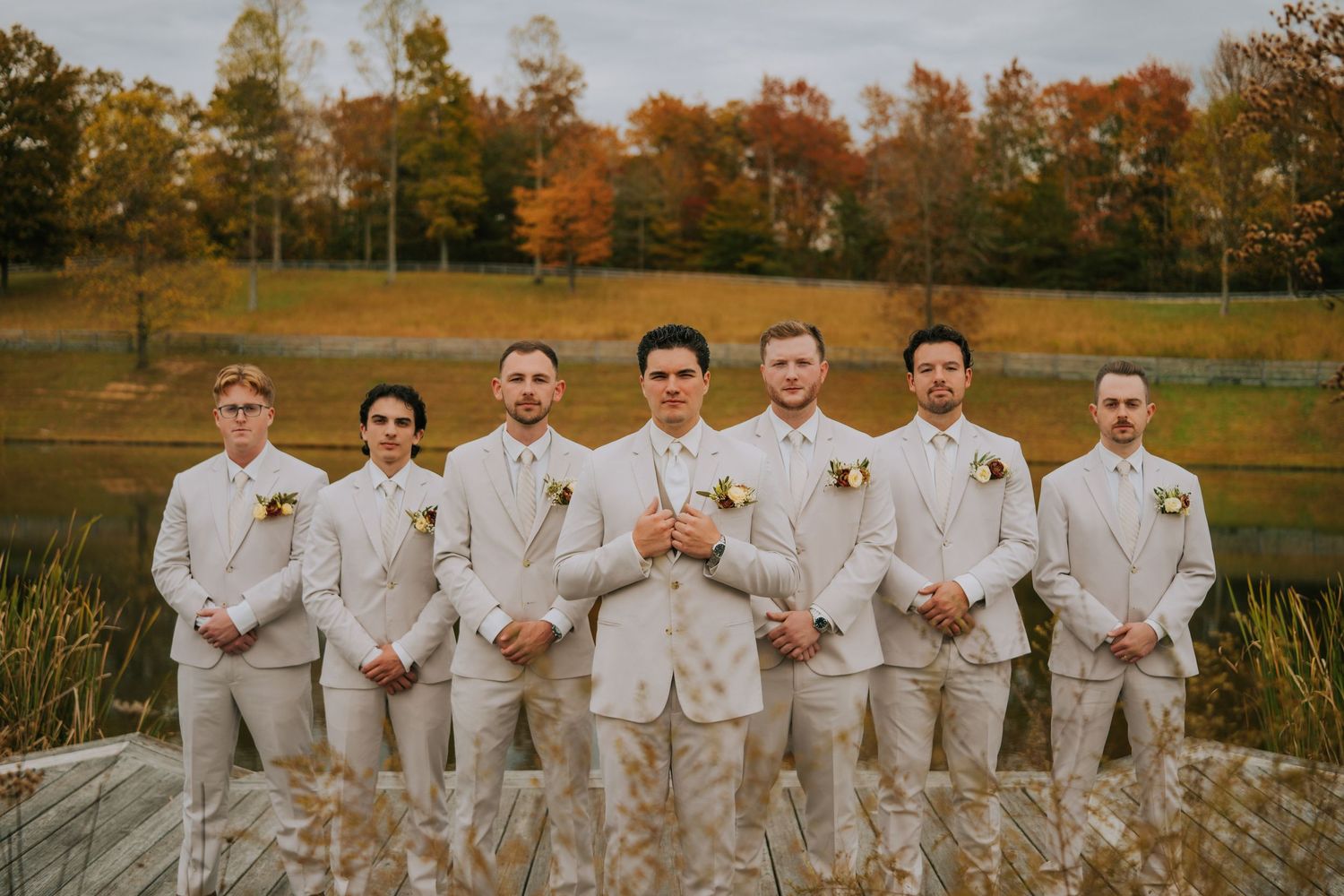 Wedding party in beige suits poses outdoors during autumn with golden foliage in the background.