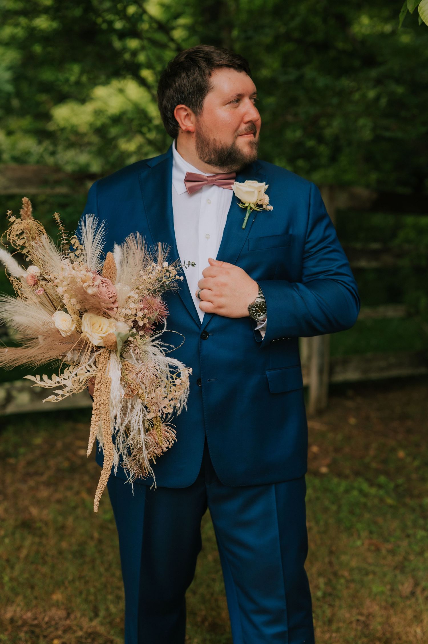 Groomsmen in navy suits hold dried pampas grass boutonnieres against a natural outdoor backdrop.