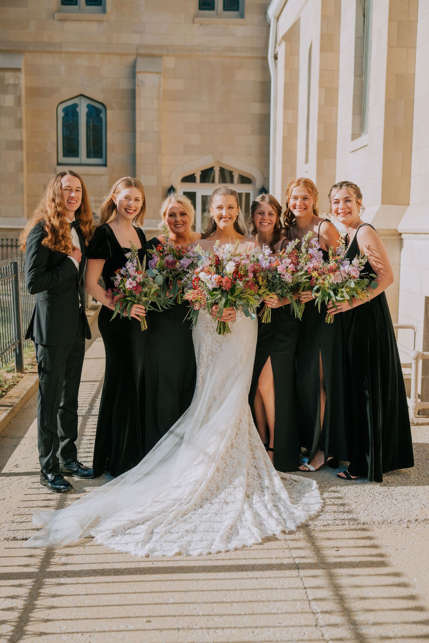 Wedding party wearing black floor-length dresses stands together holding colorful bouquets outside stone building.