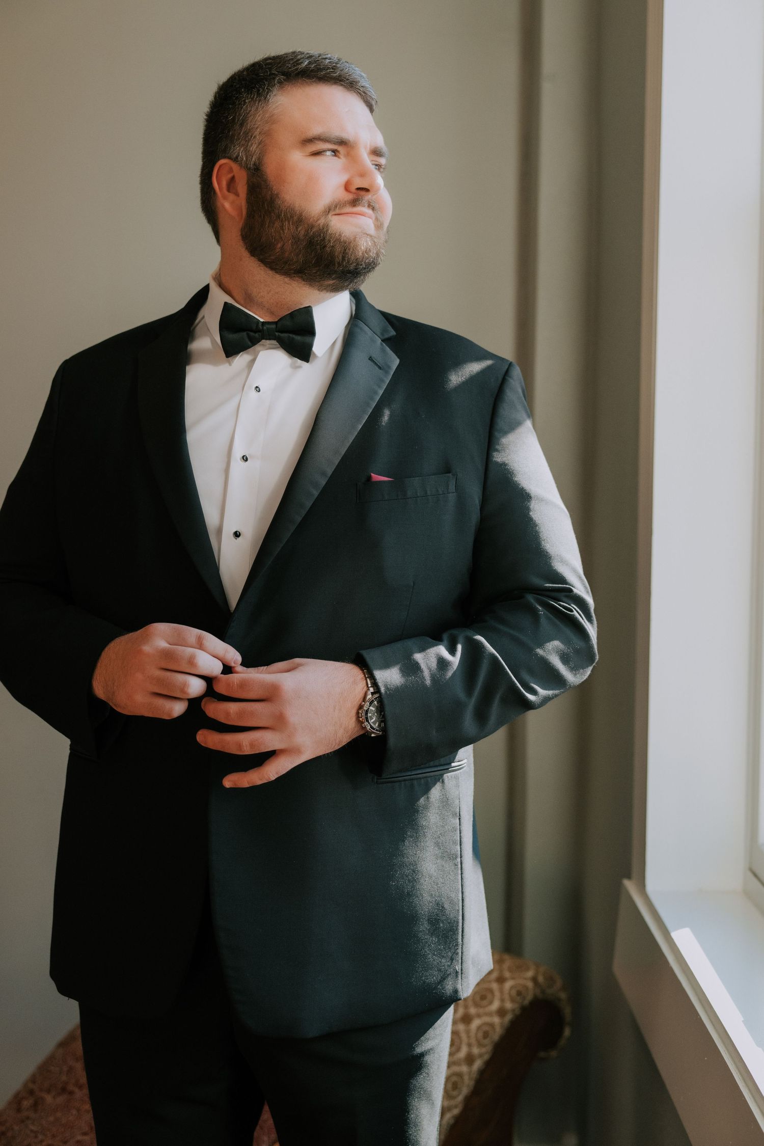 Groom in formal black tuxedo and bowtie stands thoughtfully by window in elegant pose.