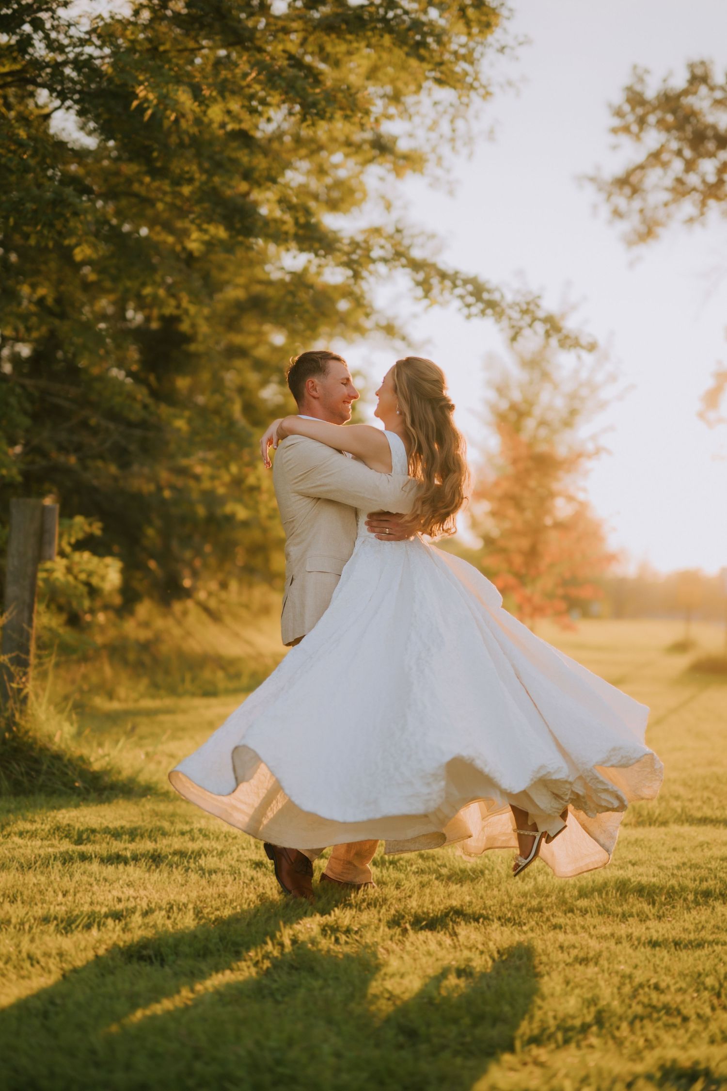 Couple dancing in golden sunset light, groom lifting bride in white flowing dress outdoors.