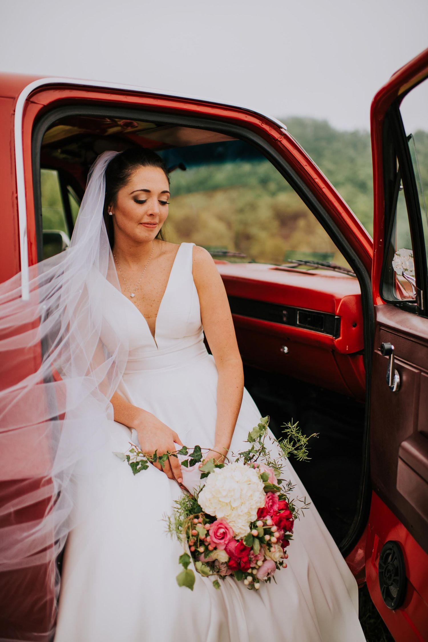 A bride in white dress poses with her bouquet in a vintage red car.