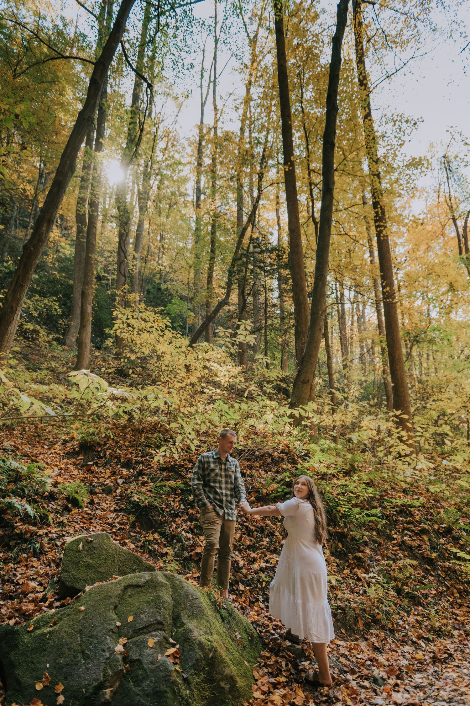 A couple poses on fallen autumn leaves among large boulders in a sunlit forest.