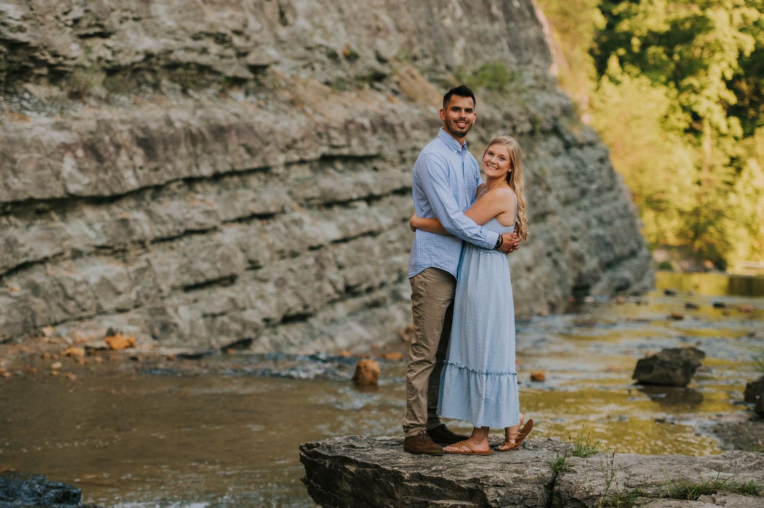A couple embraces while standing on a rock formation near flowing water during golden hour.