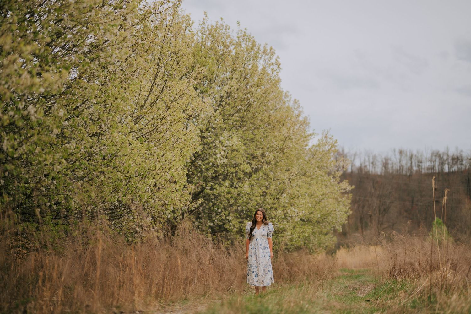 Woman in floral dress standing in distance among tall grass and trees.
