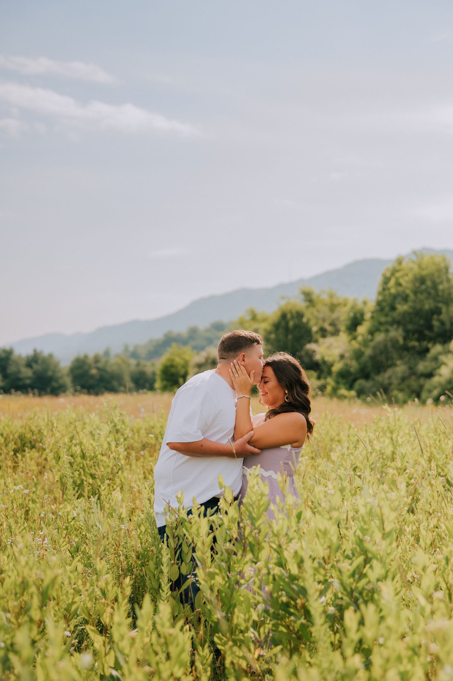 Two people share an intimate moment in a sunny wildflower field with mountains in the distance.