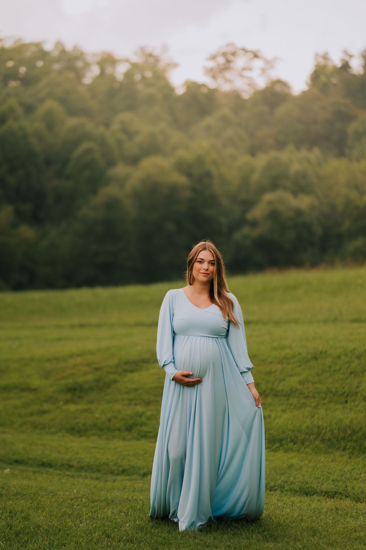 A maternity session featuring a woman in a light blue maxi dress posing in a green field.
