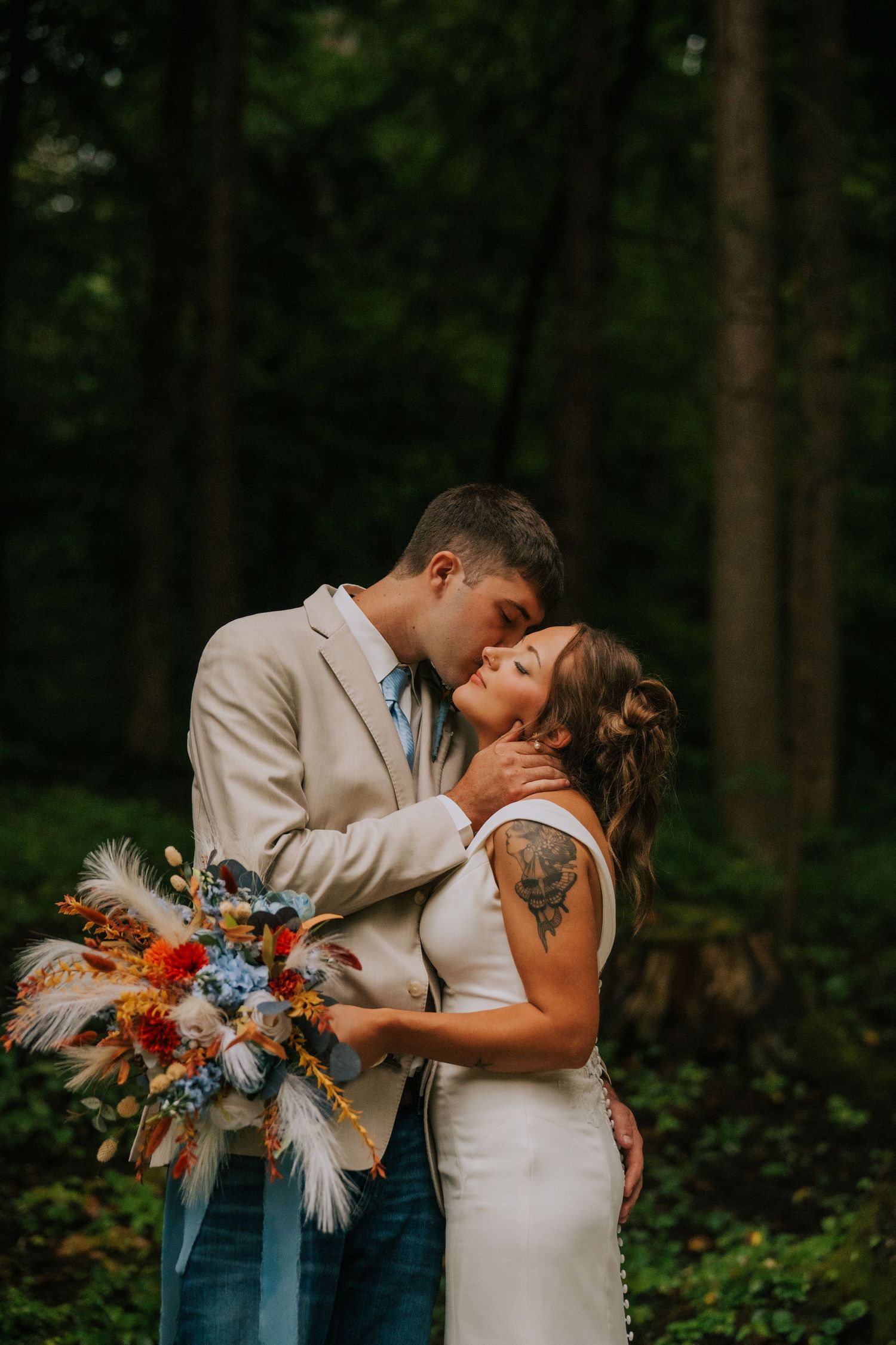 A couple shares a romantic moment in a forest setting with a colorful floral bouquet featuring pampas grass.