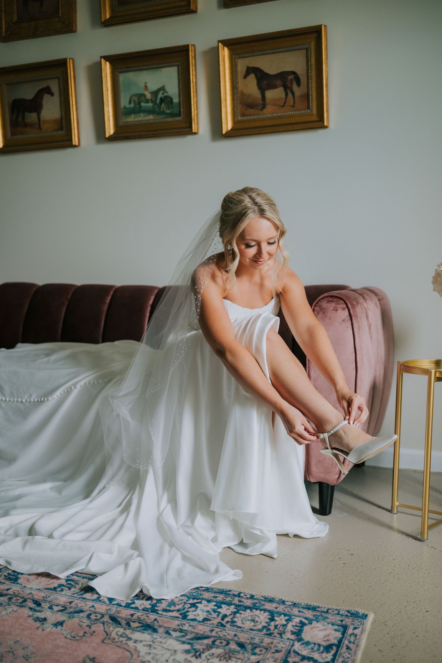 Bride putting on wedding shoes while sitting on pink couch beneath framed horse paintings.