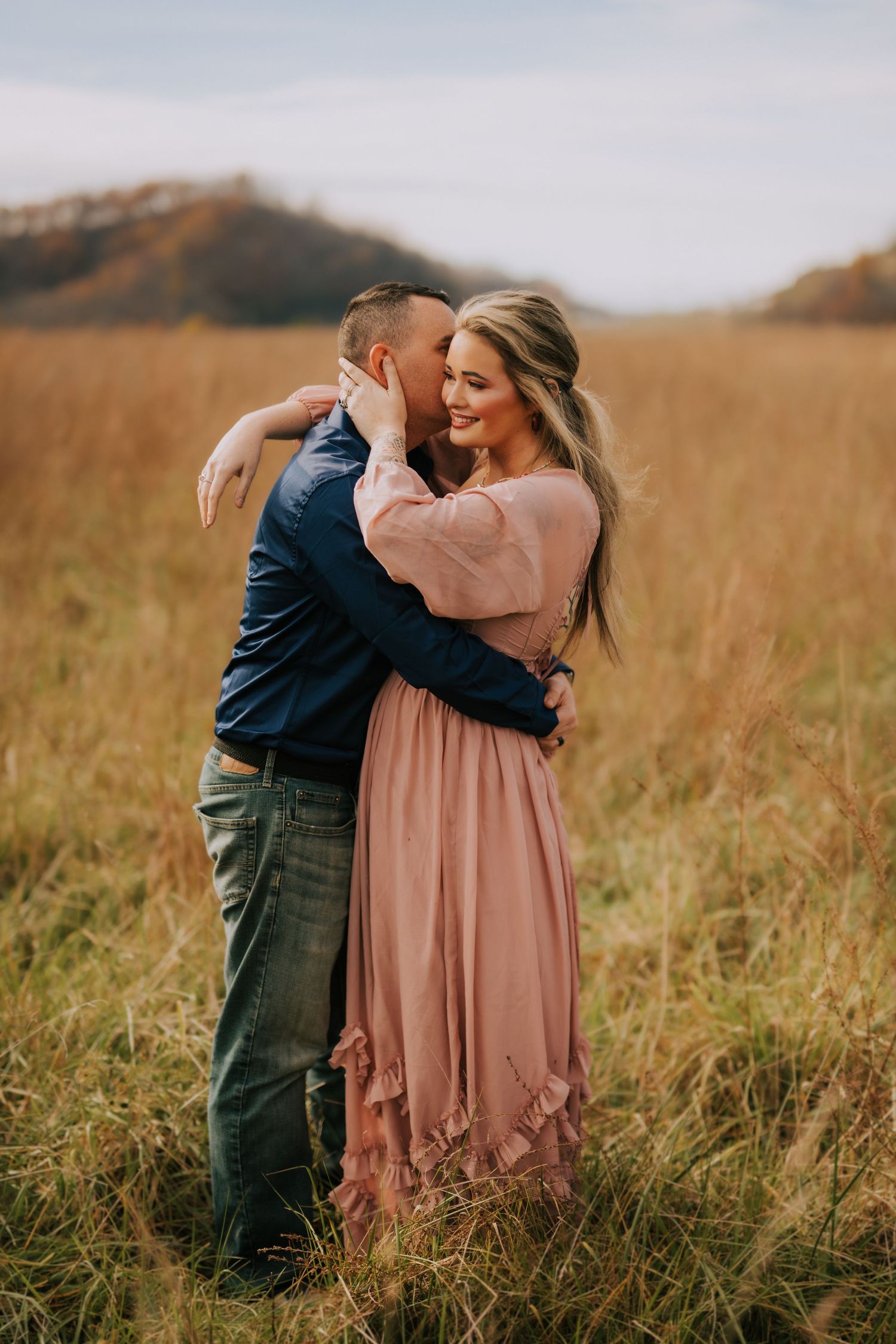 Romantic couple embraces in a golden field during sunset wearing pink dress and navy blue outfit.