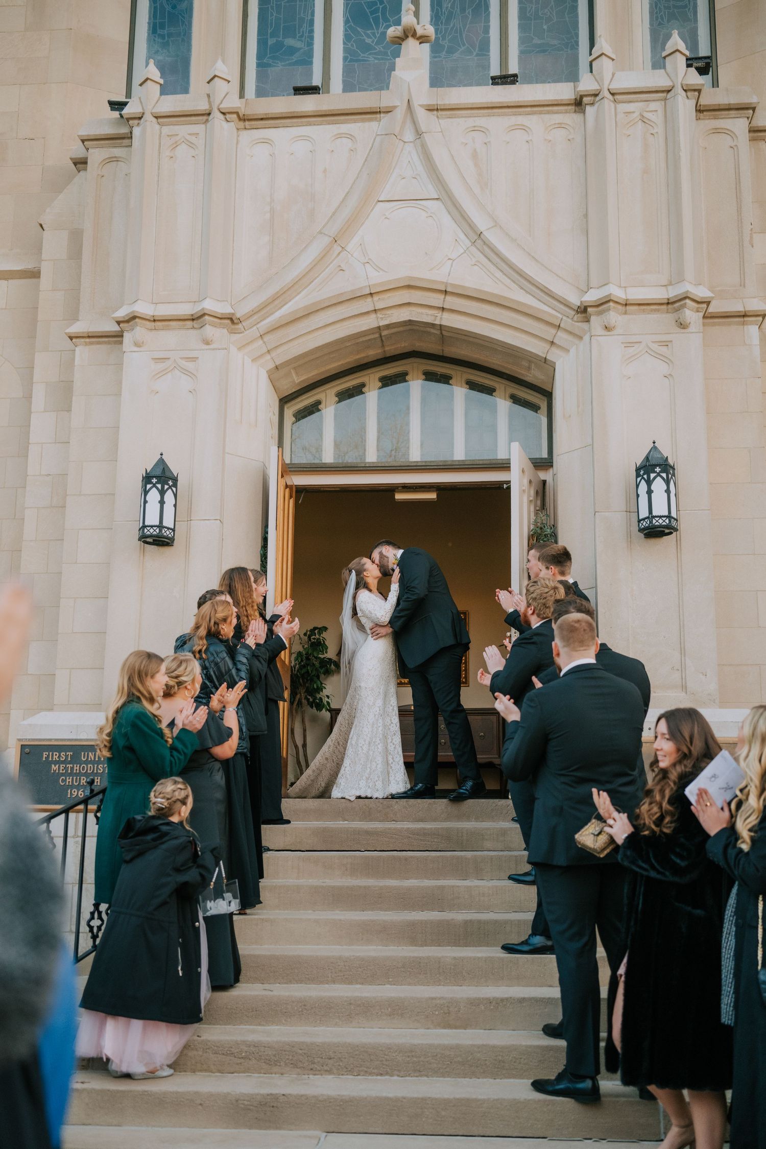 Wedding ceremony guests in formal attire gather on church steps as couple exits through Gothic archway entrance.