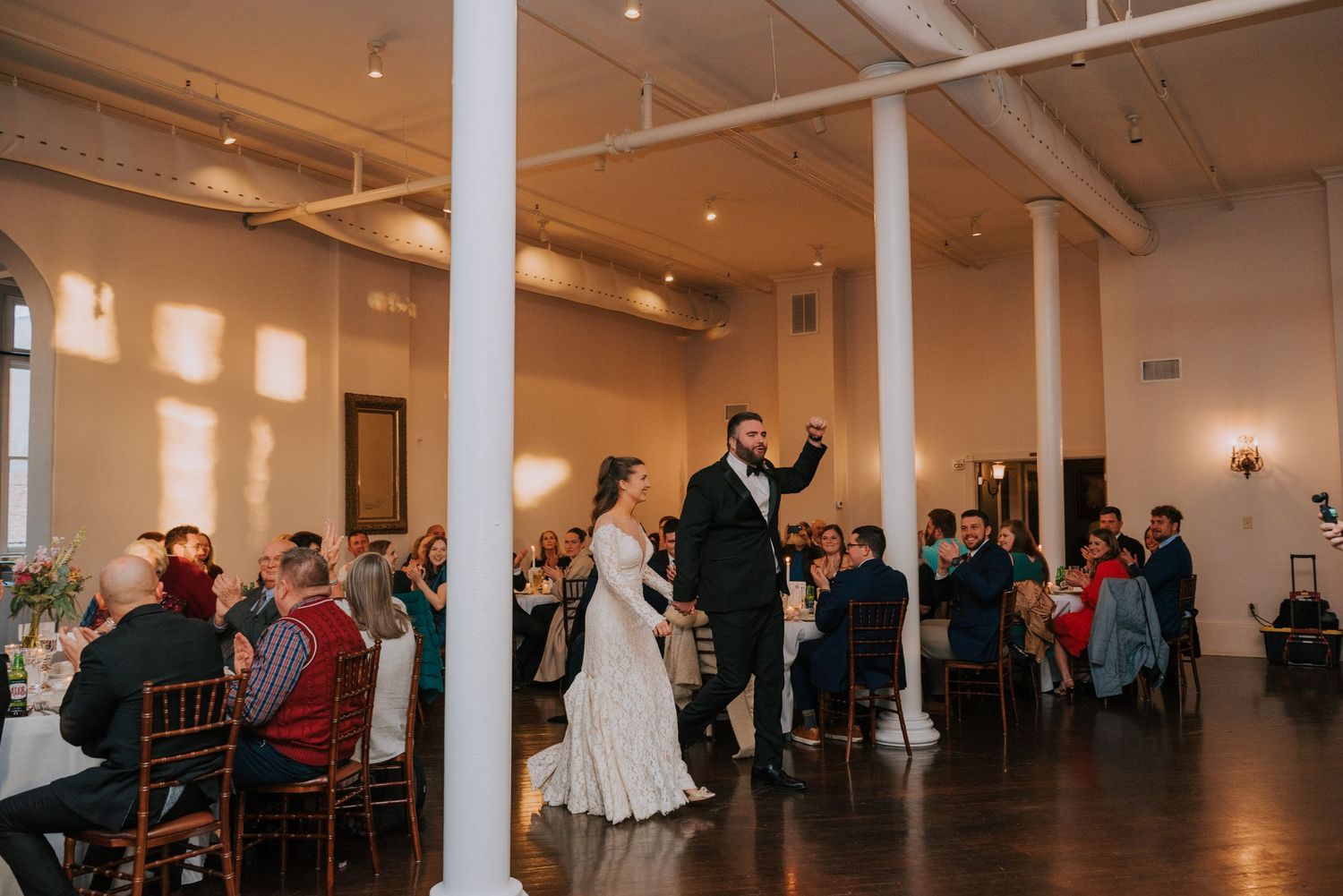 Wedding reception guests watch couple make a toast in an elegant room with tall columns and warm lighting.