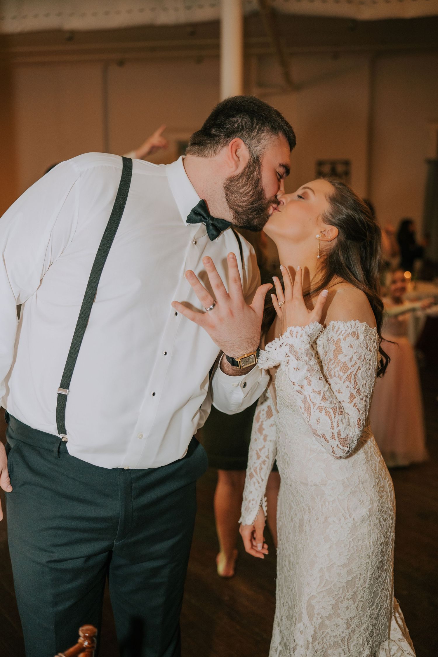 Newlyweds share romantic kiss at wedding reception with bride in lace long sleeve dress and groom in suspenders.
