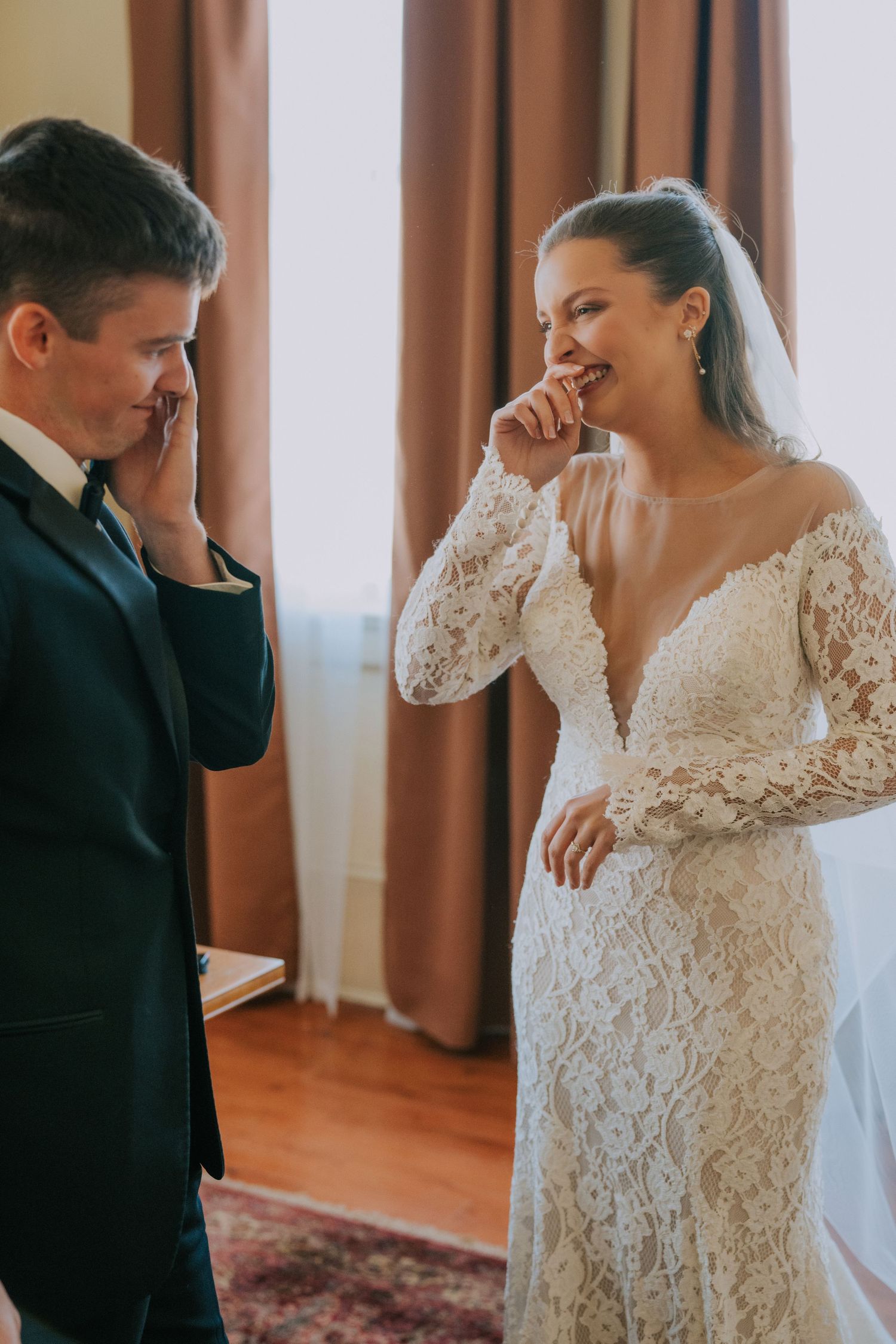 Emotional first look moment between couple on their wedding day inside room with lace gown and dark suit.