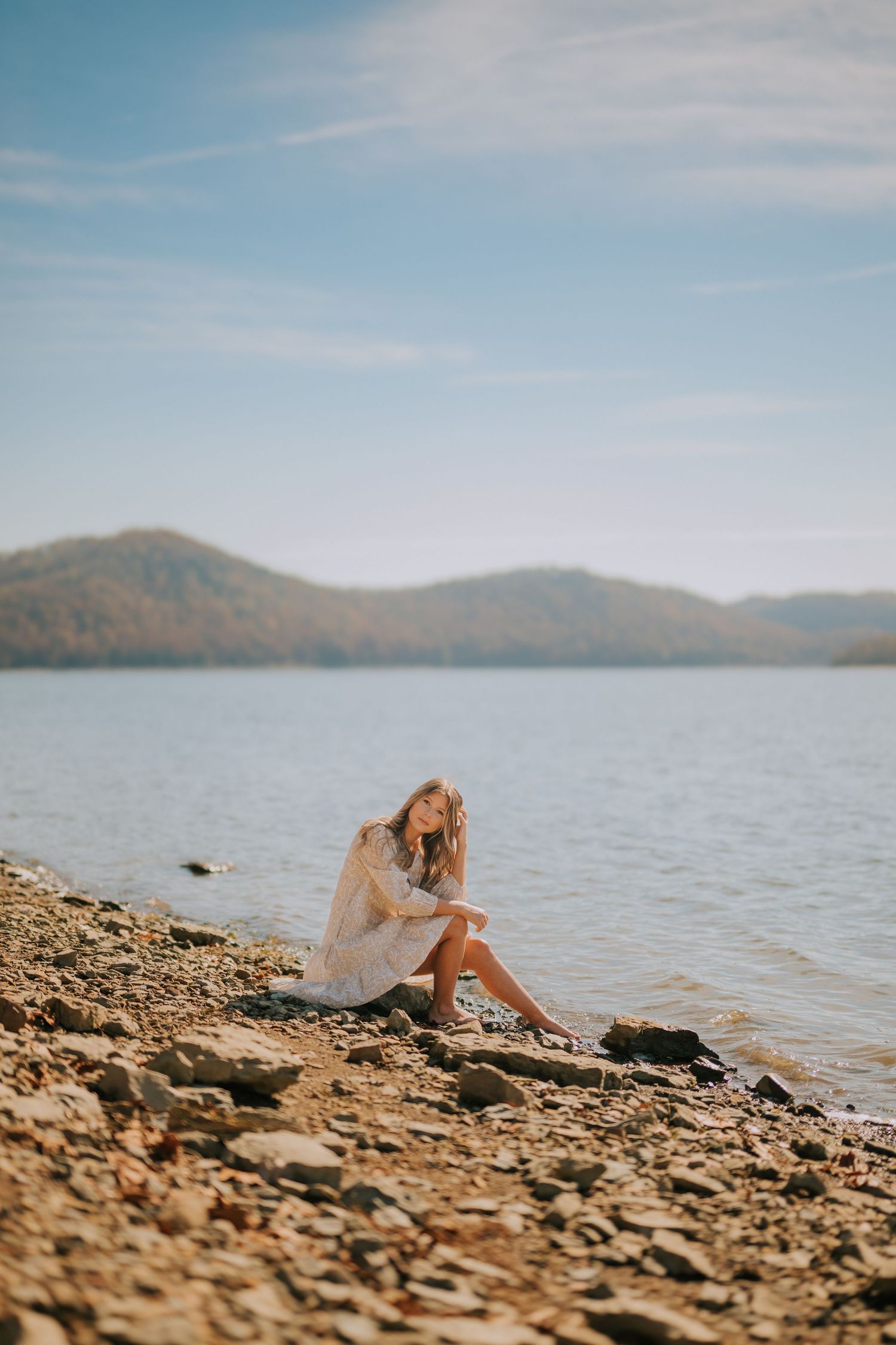 Person in white dress sits on rocky shoreline overlooking serene mountain lake.