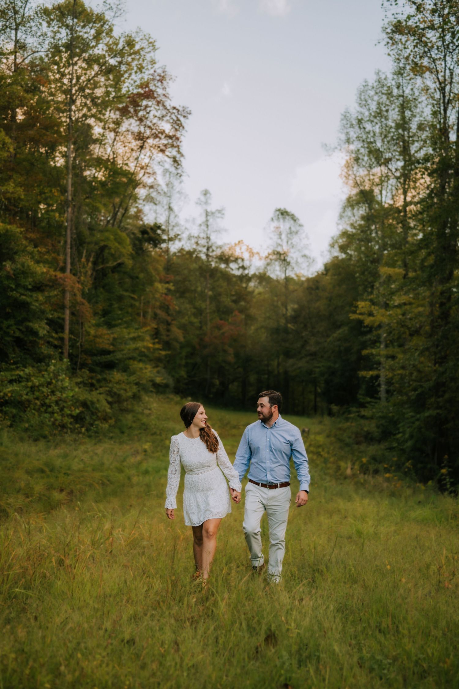 Couple walks hand in hand through a serene meadow surrounded by trees.