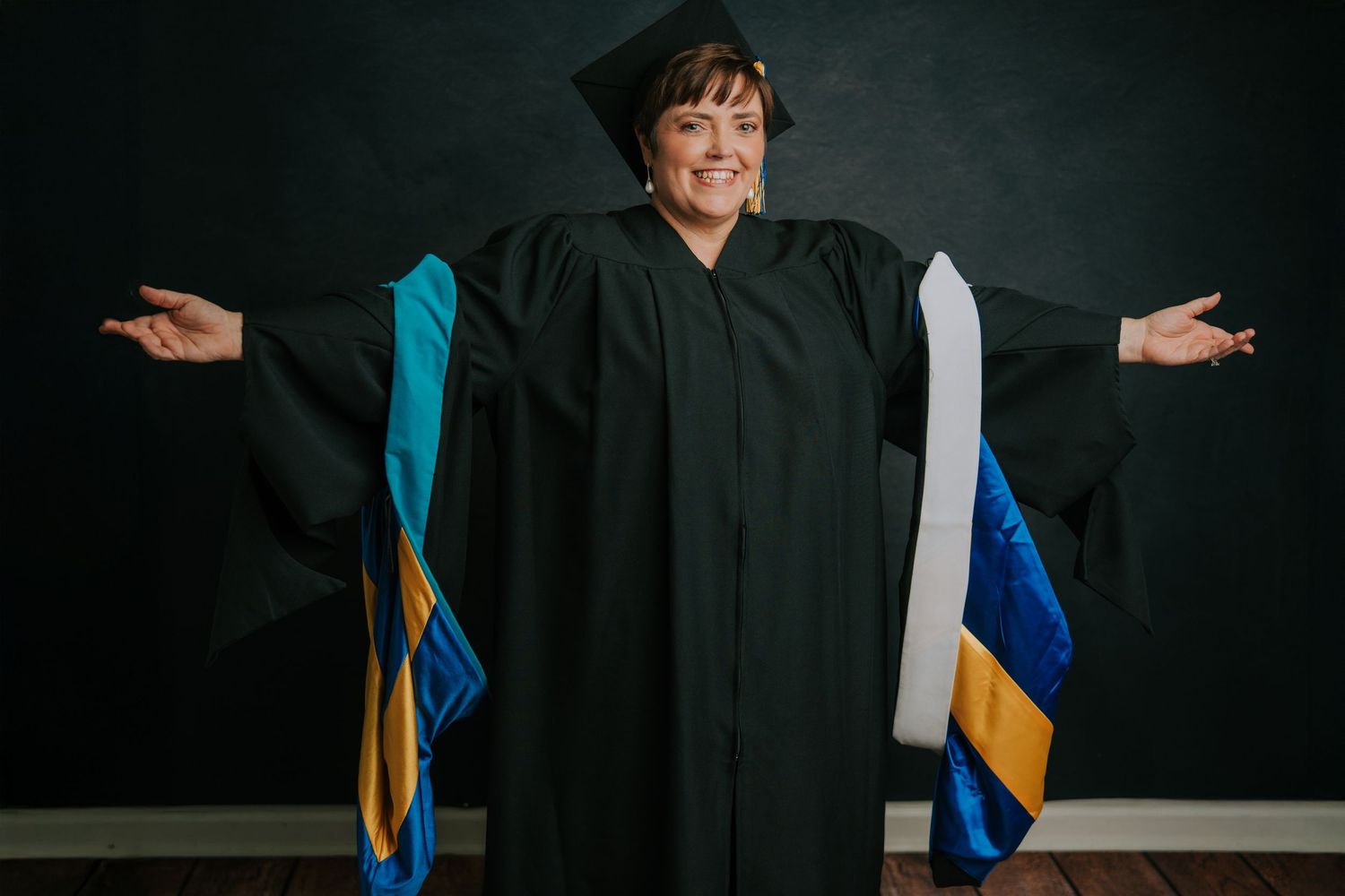 Graduate in traditional academic regalia gestures with outstretched arms against dark backdrop.