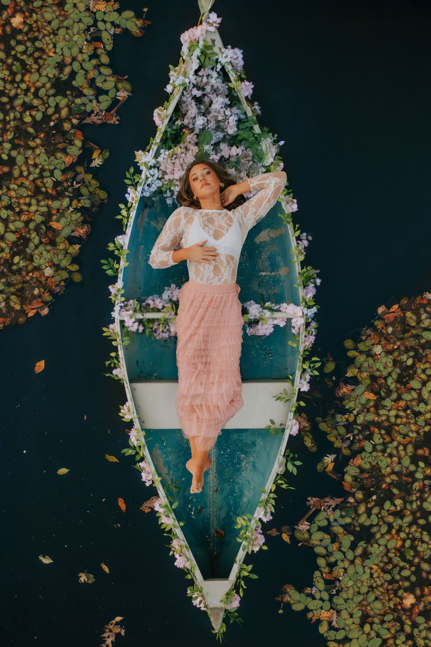 Dreamy portrait series in pink dress lying on floral-adorned swing against dark background.